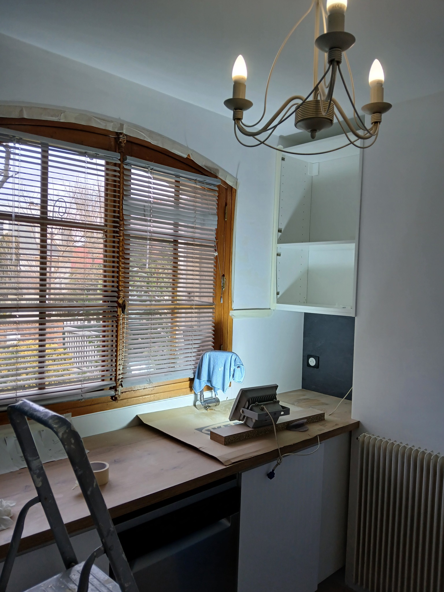 Wnętrze w trakcie prac: drewniany blat zabezpieczony papierem, drabina, lampa budowlana, wisząca szafka kuchenna, okno z żaluzjami. Przygotowania do montażu.