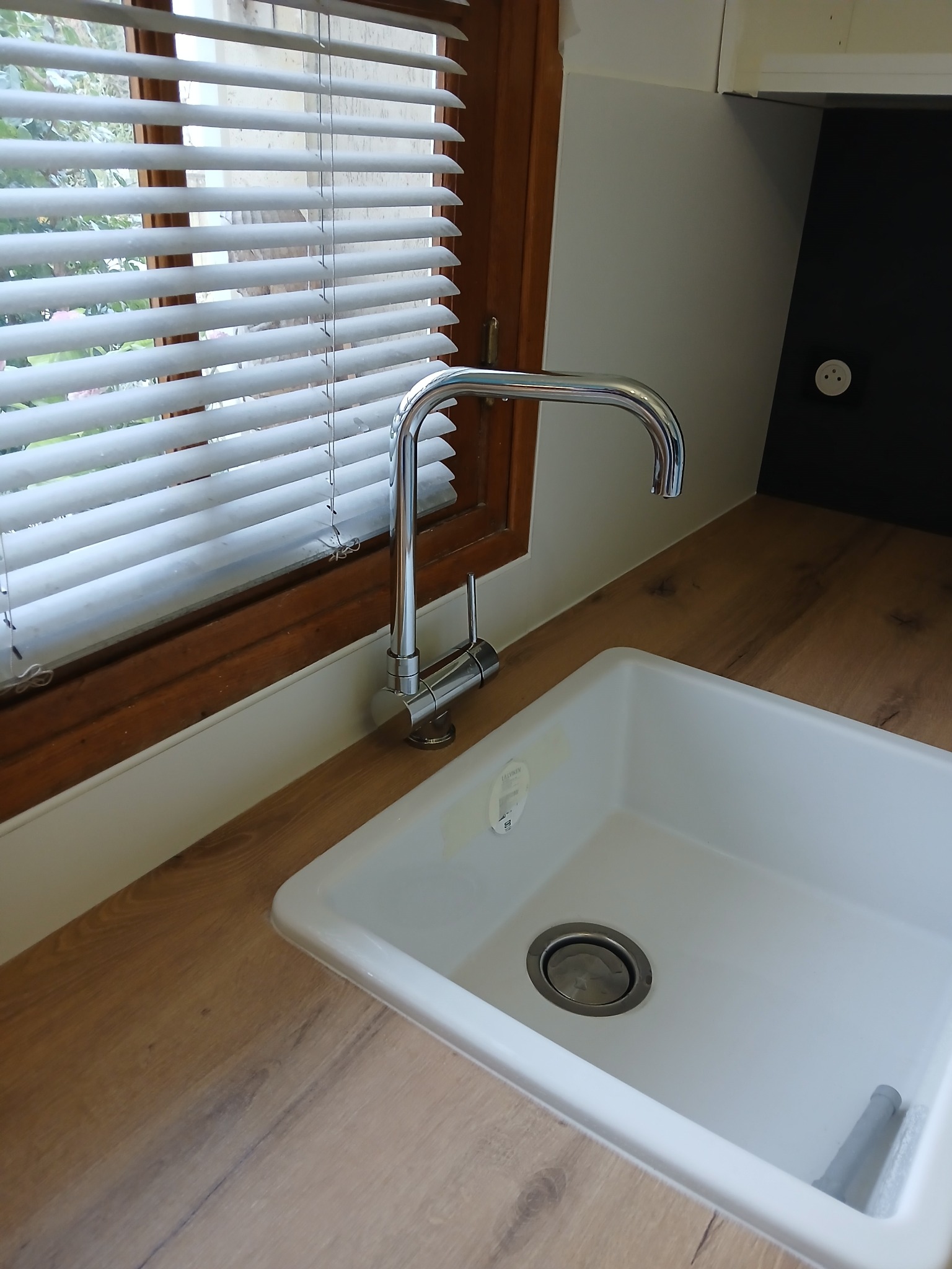 Biały zlew z chromowaną baterią przy oknie z żaluzjami, na blacie imitującym drewno. Nowoczesna aranżacja kuchni po remoncie, jasne wnętrze.