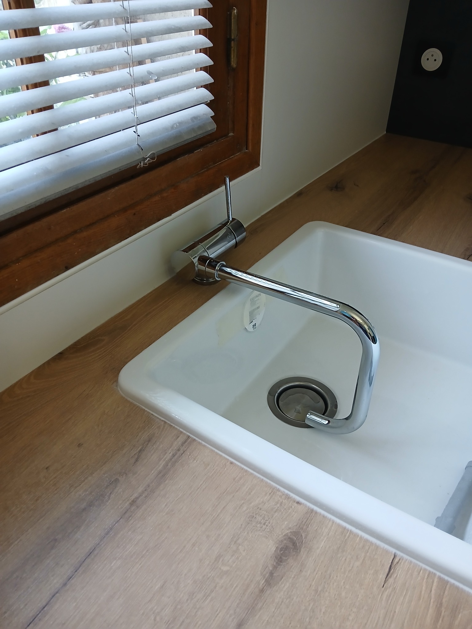 Biały zlew z chromowaną baterią na blacie imitującym drewno, przy oknie z żaluzjami. Widoczny fragment ściany z gniazdkiem elektrycznym. Ujęcie z góry.