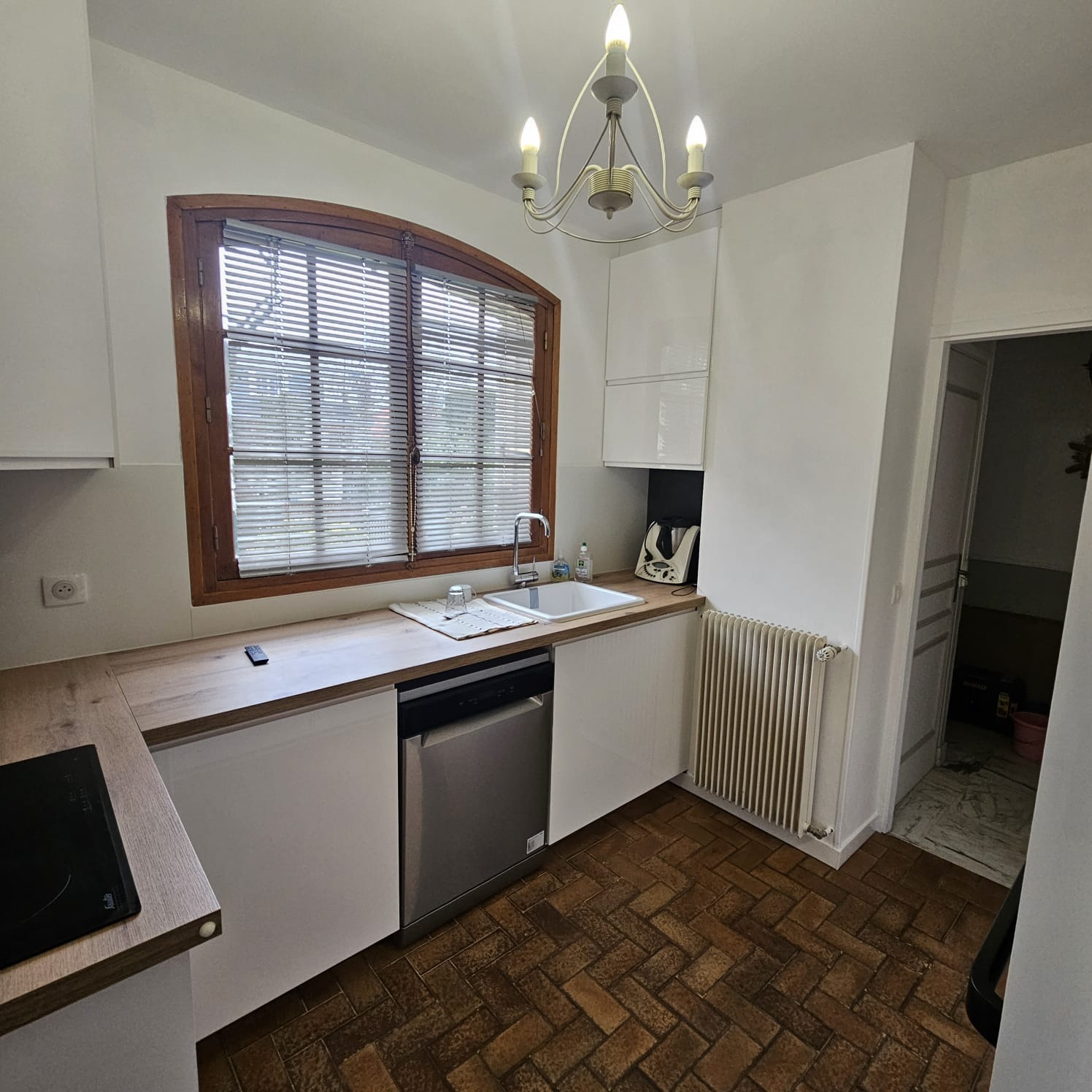 Jasna kuchnia z drewnianą podłogą w jodełkę, białymi szafkami i oknem z żaluzjami. Widoczny fragment płyty grzewczej i grzejnik. Ciepłe światło żyrandola.