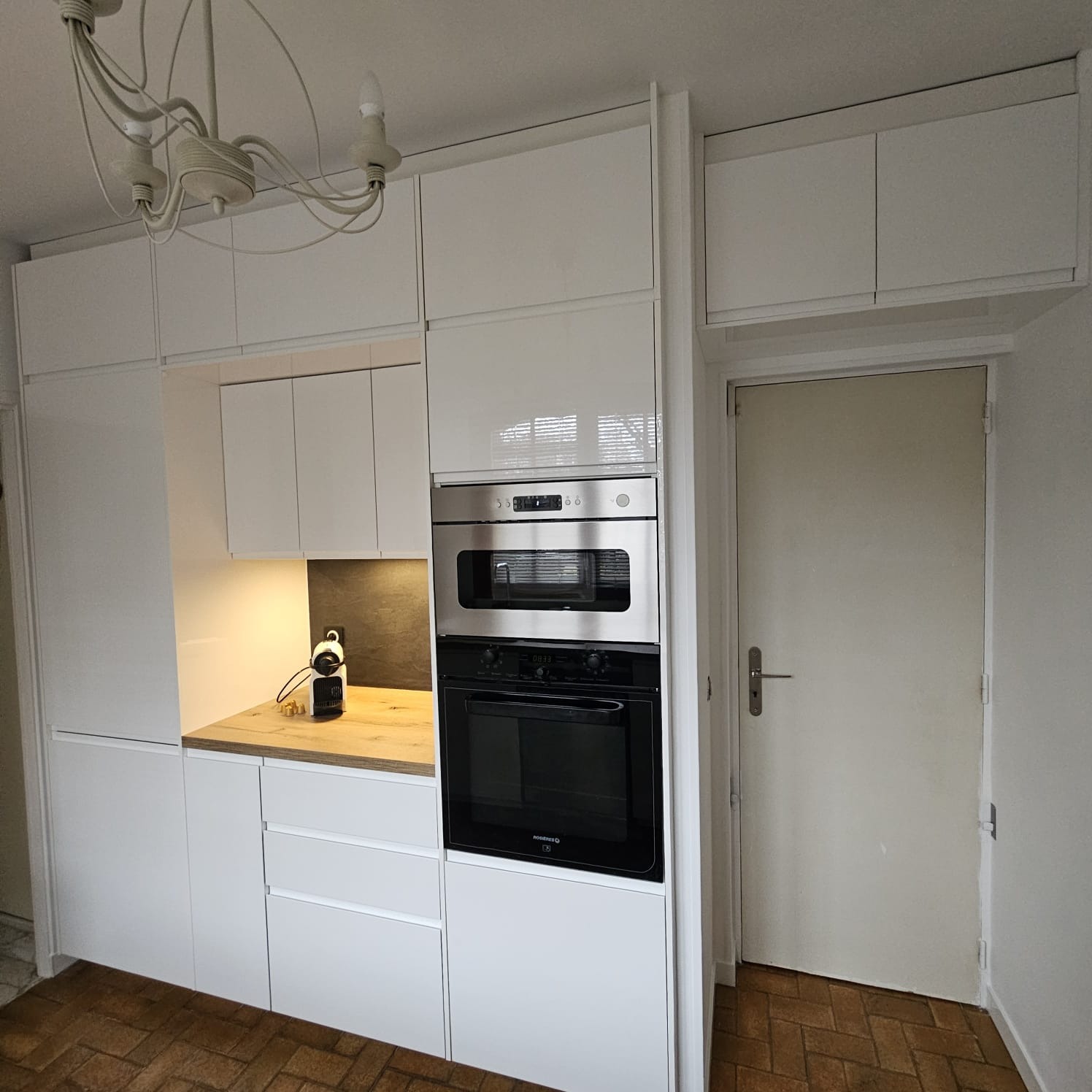 Biała zabudowa kuchenna z piekarnikiem i mikrofalówką, drewniany blat, parkiet w jodełkę, minimalistyczny żyrandol. Nowoczesne wykończenie wnętrza kuchni.