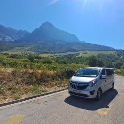 JS RENT Sebastian Jurczak - Srebrny bus Opel Vivaro zaparkowany na poboczu drogi z widokiem na góry i zielone wzgórza w słoneczny dzień. Widoczne inne pojazdy na drodze.