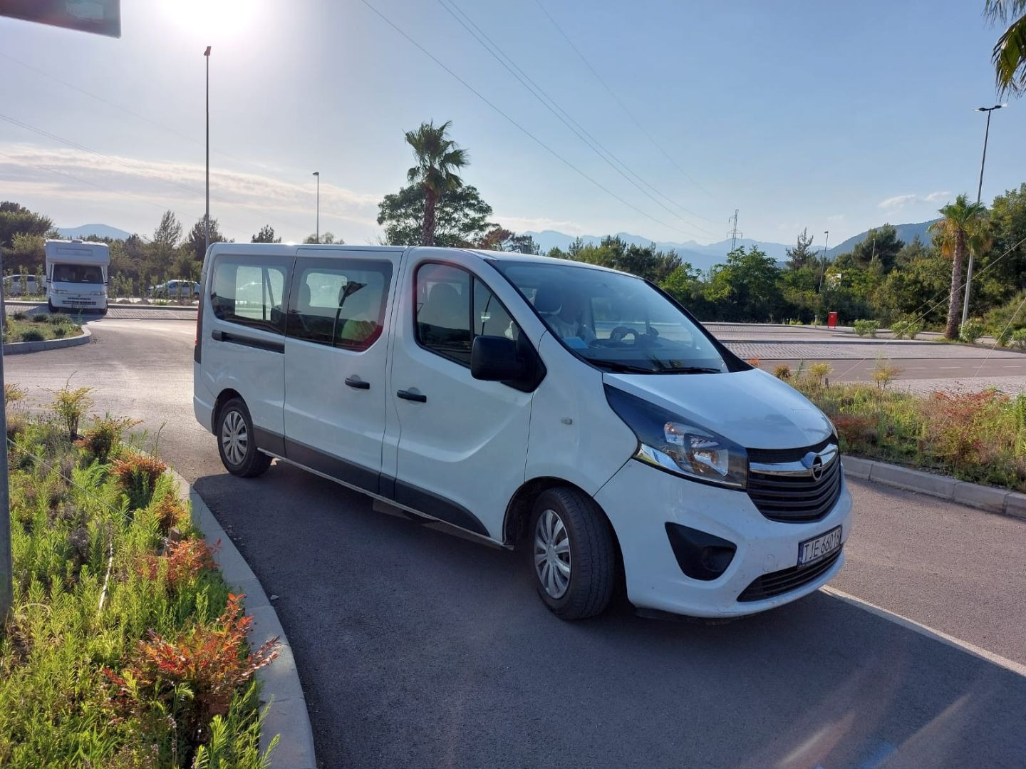 Biały Opel Vivaro na parkingu w słoneczny dzień. Widoczne drzewa, góry w tle i kamper. Tablica rejestracyjna TJE 660.