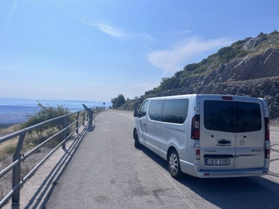 Srebrny bus Opel Vivaro z polską rejestracją na tle malowniczego krajobrazu z widokiem na morze i góry. Pojazd zaparkowany na poboczu drogi.