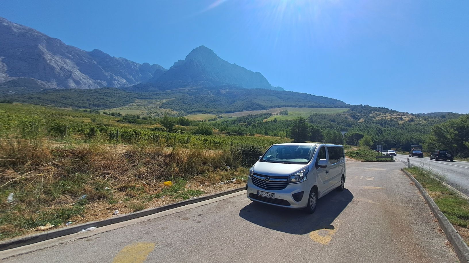 Srebrny bus Opel Vivaro zaparkowany na poboczu drogi z widokiem na góry i zielone wzgórza w słoneczny dzień. Widoczne inne pojazdy na drodze.