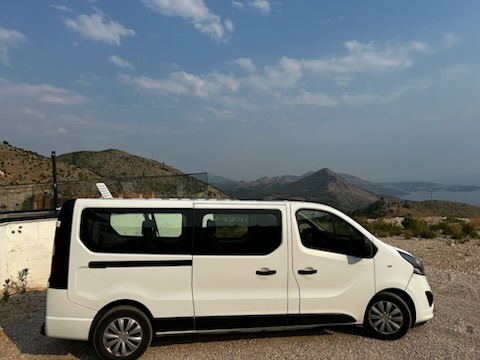 Biały bus na tle górskiego krajobrazu z błękitnym niebem. Pojazd zaparkowany na żwirowej drodze, widok z boku, podkreślający przestronność i komfort podróży.
