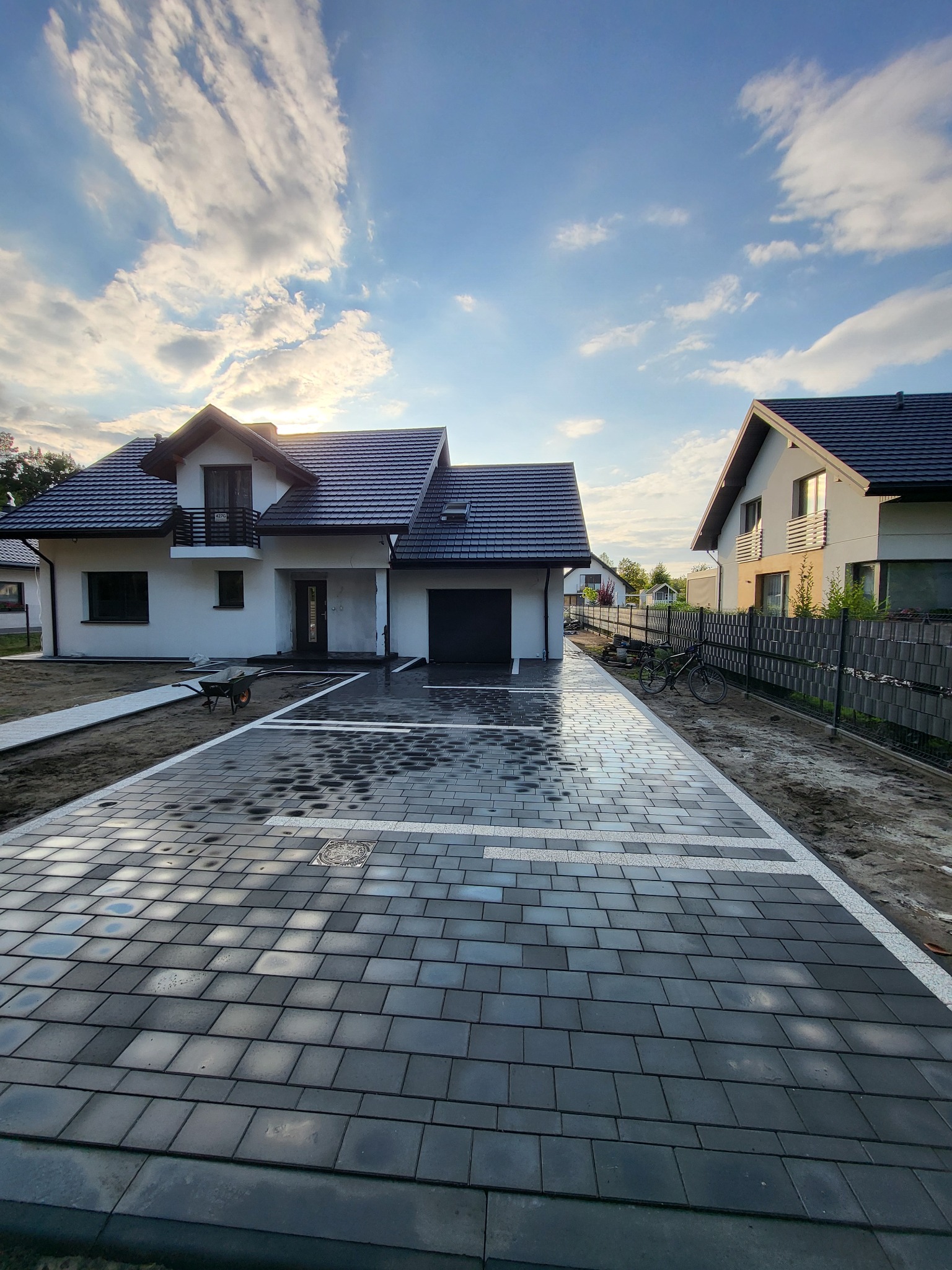 Nowoczesny podjazd z szarej kostki brukowej, ułożonej w geometryczny wzór z białymi akcentami, prowadzący do domu z garażem. Widoczne niebo z chmurami.