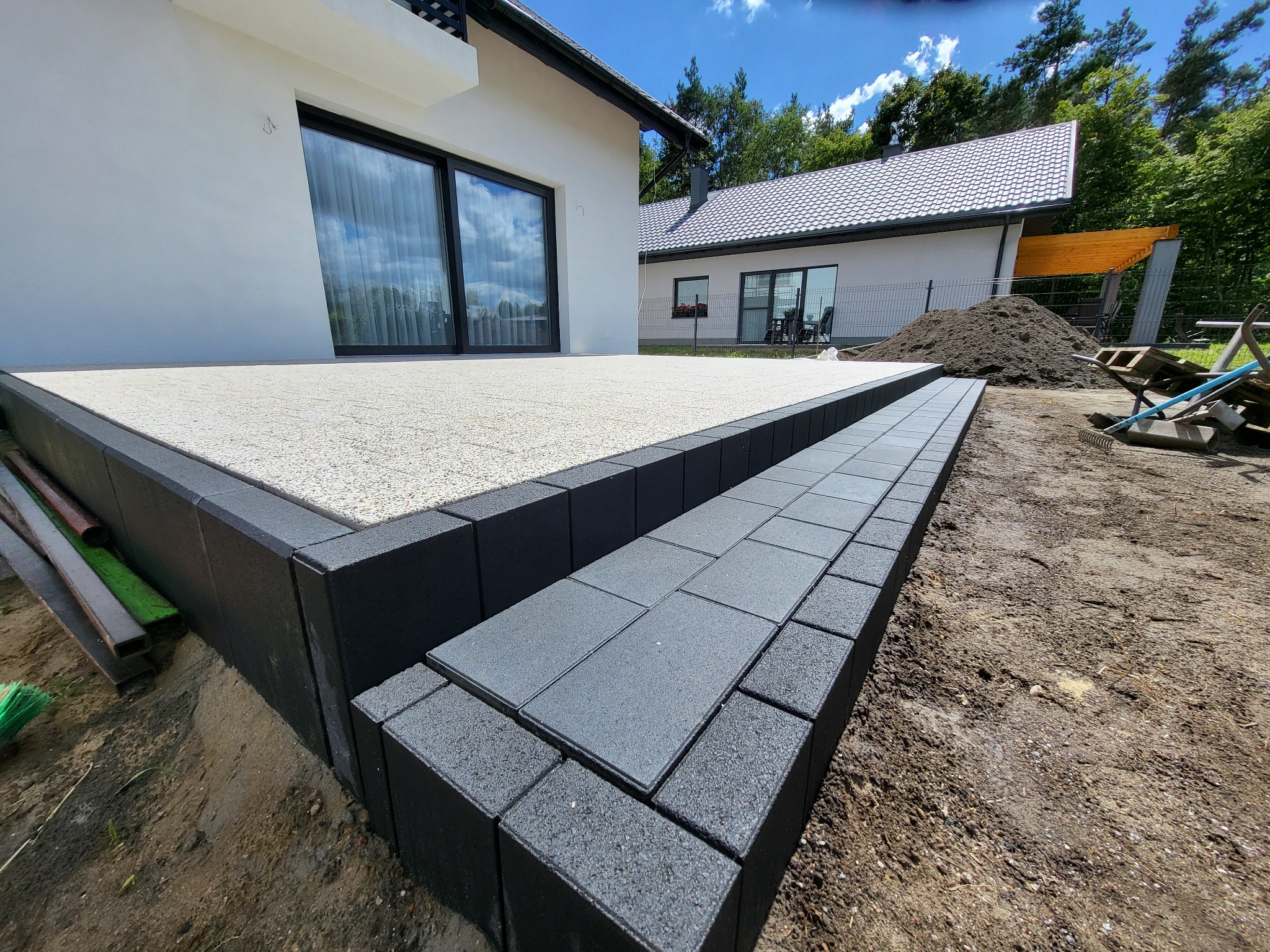 Wykończony taras z jasną nawierzchnią i schodami z ciemnej kostki brukowej, obrzeża z czarnych elementów. W tle dom z dużymi oknami i drugi budynek.