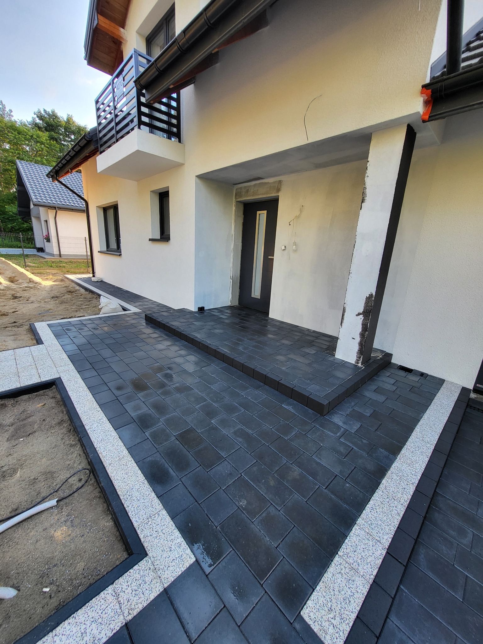 Nowoczesna posesja z ciemną kostką brukową obramowaną jasnym kamieniem, prowadząca do wejścia domu. Widoczny fragment ogrodu i elewacja budynku z balkonem.