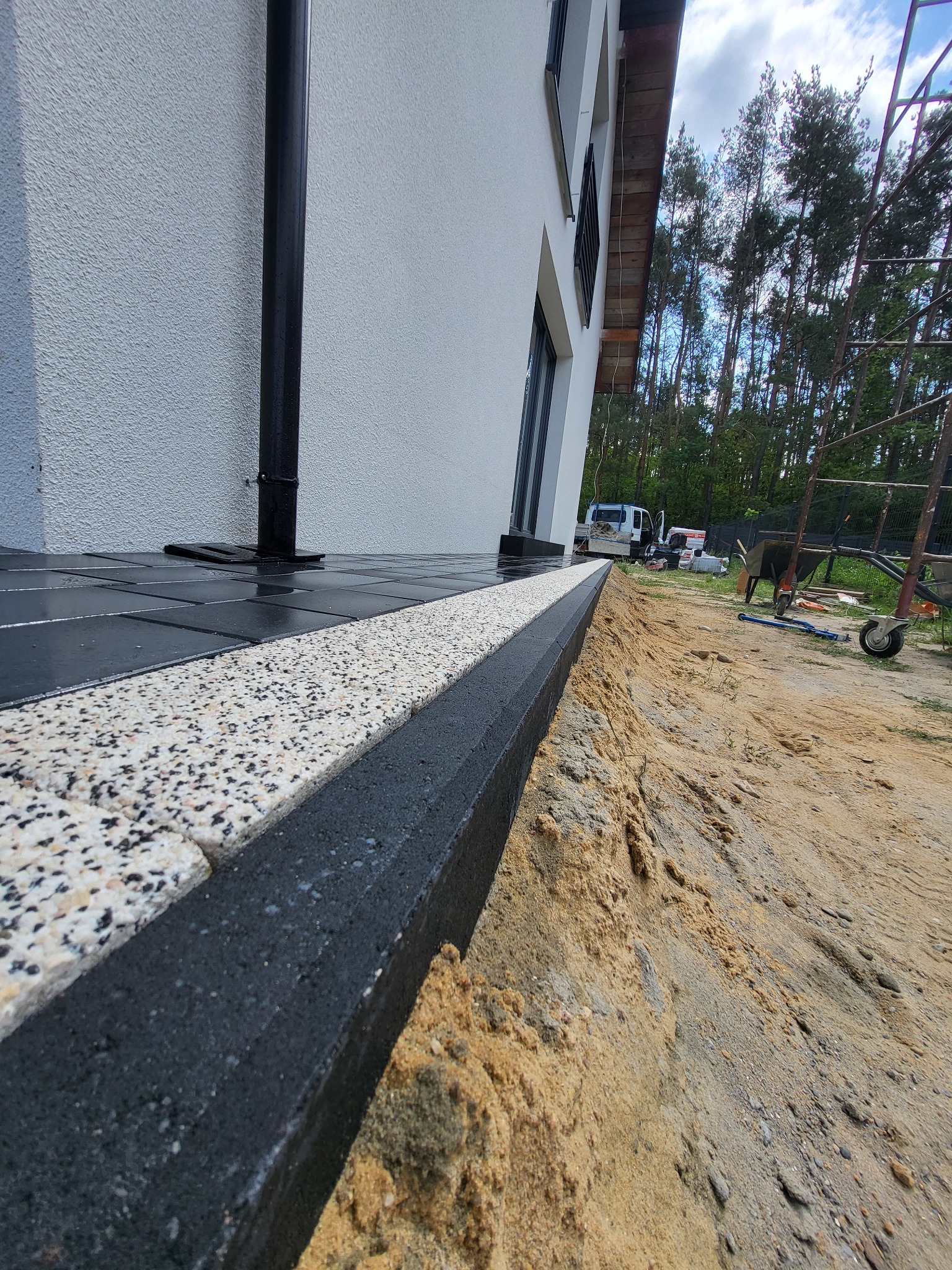 Wykończony fragment ścieżki z kostki brukowej przy budynku, z widocznym obrzeżem i przygotowanym podłożem. Ujęcie z niskiej perspektywy podkreśla detale.