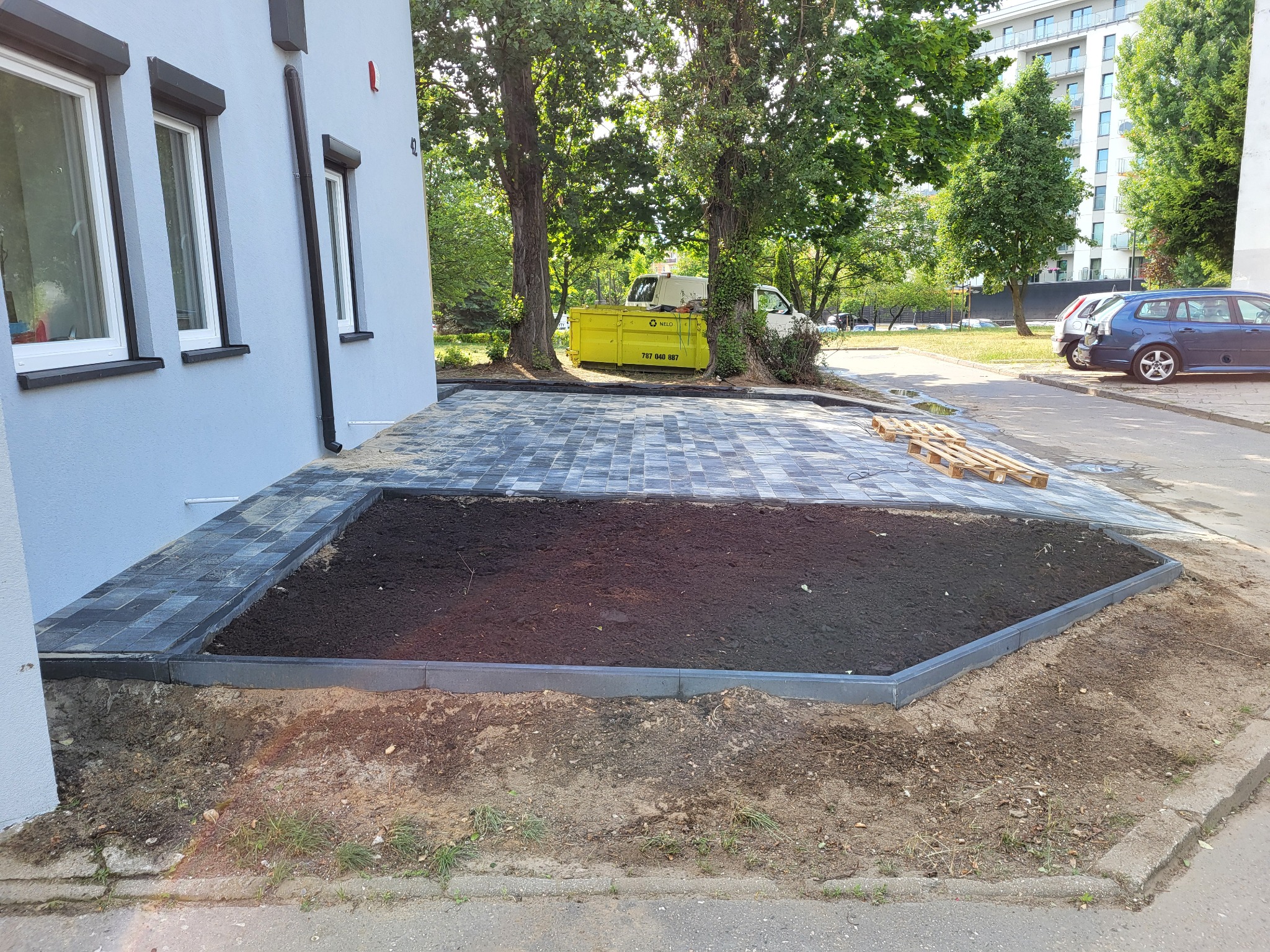 Ułożona kostka brukowa w szaro-czarnych odcieniach, obramowana rabata z ziemią, widok z boku budynku. W tle zieleń drzew i zaparkowane samochody.