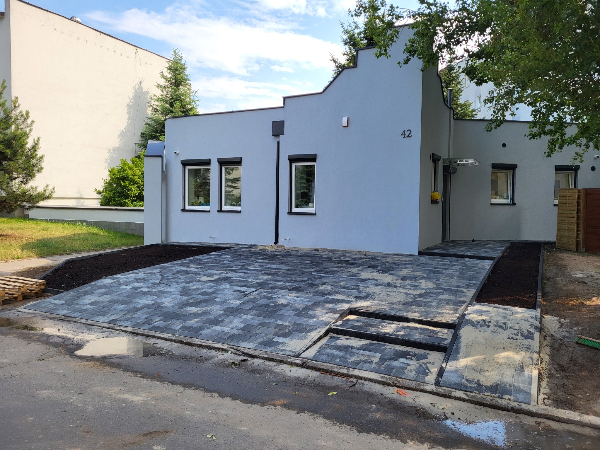 Nowoczesny podjazd z szarej kostki brukowej przed budynkiem z numerem 42. Widoczne fragmenty niezakończonych prac i paleta. Ujęcie z poziomu gruntu.