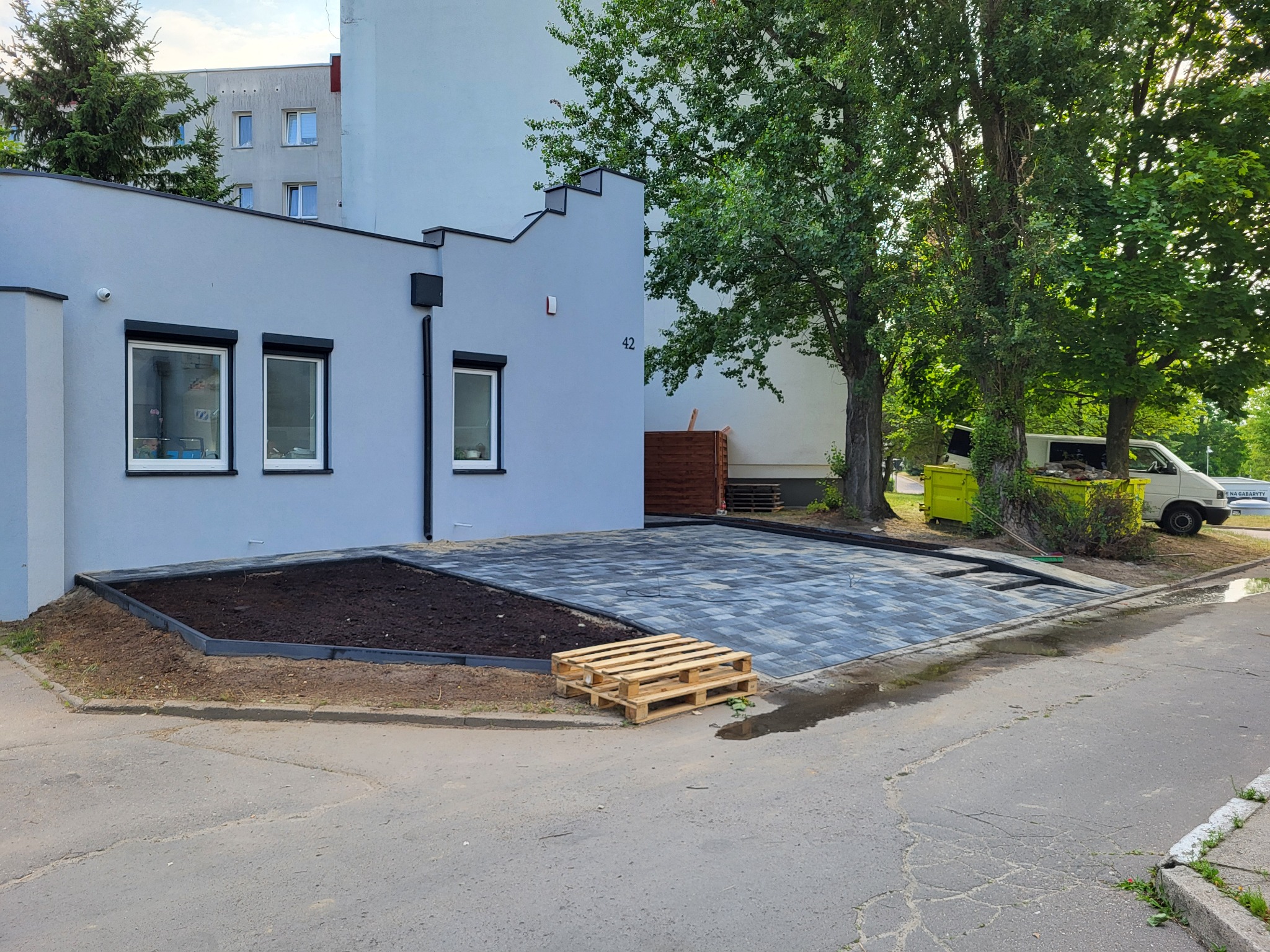 Częściowo ułożona, szara kostka brukowa na podjeździe przed budynkiem z numerem 42. Obok przygotowane miejsce pod rabatę. Paleta drewniana na pierwszym planie.