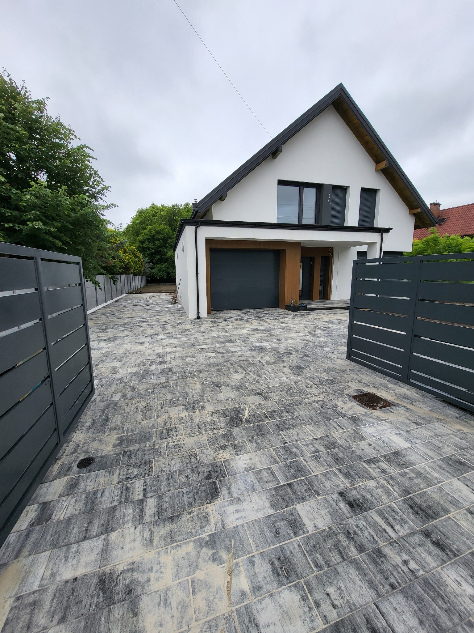 Nowoczesny podjazd z szarej kostki brukowej prowadzący do domu z garażem i szarą bramą. Widoczne otwarte, metalowe ogrodzenie i zieleń w tle.