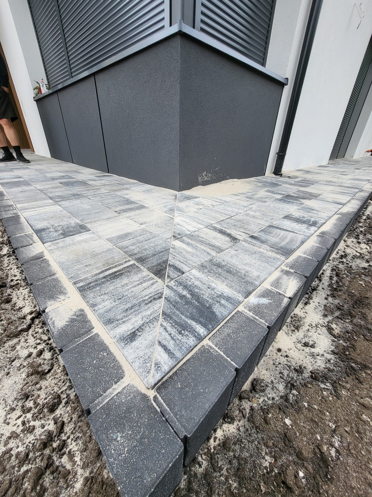 Świeżo ułożona, szara kostka brukowa tworząca ścieżkę przy nowoczesnym budynku z ciemnymi elementami elewacji. Widoczne fugi wypełnione piaskiem i fragment nogi osoby.