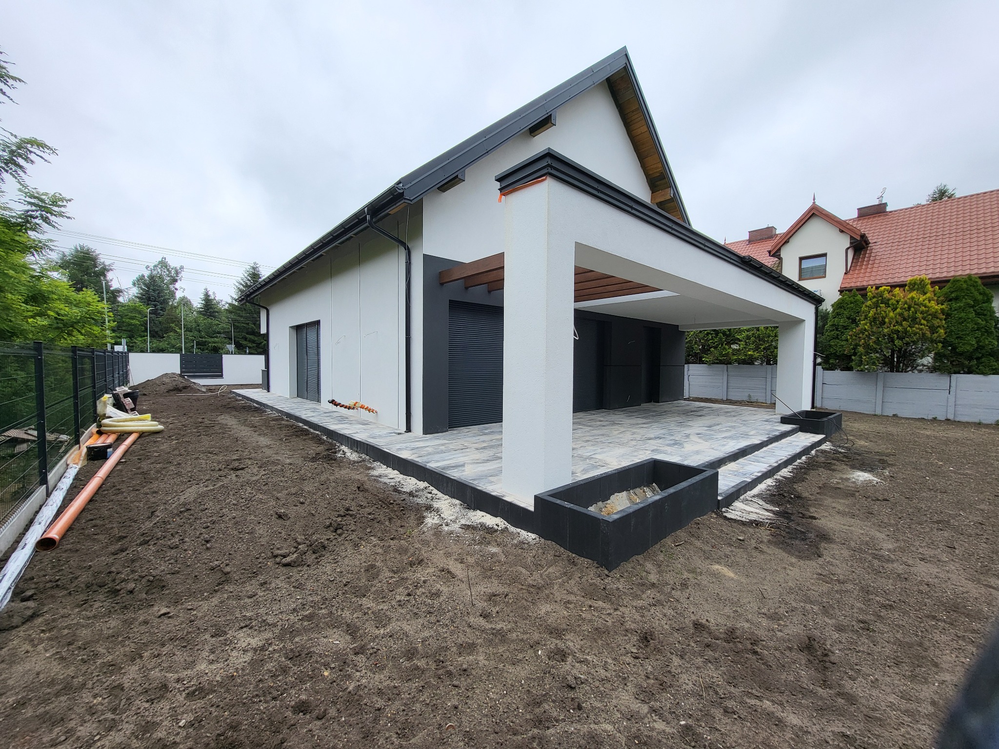 Nowoczesny taras z kostki brukowej w szarościach, przylegający do białego domu z czarnymi akcentami. Widoczne przygotowania terenu pod dalsze prace.