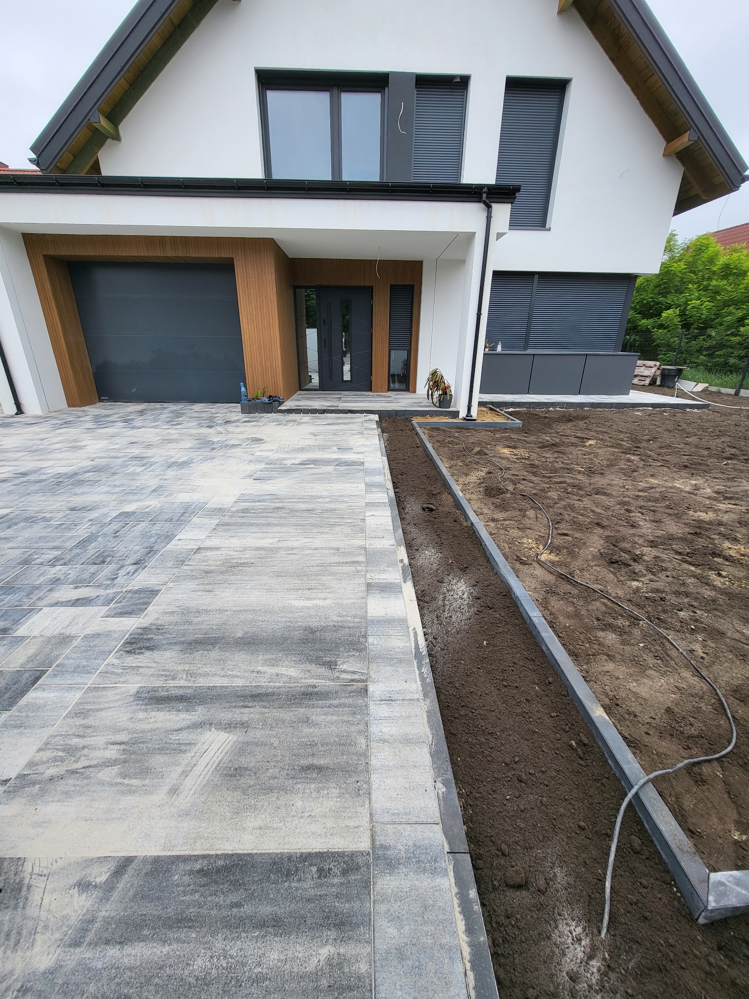 Nowoczesny podjazd z szarej kostki brukowej prowadzący do domu z garażem i drewnianymi akcentami. Częściowo przygotowany teren pod dalsze prace.