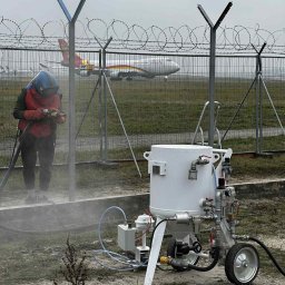 DECART sp. z. o o. - Pracownik w kombinezonie ochronnym czyści metalowy słup za pomocą piaskarki mobilnej na tle lotniska i samolotu. Widoczna mgła piasku.