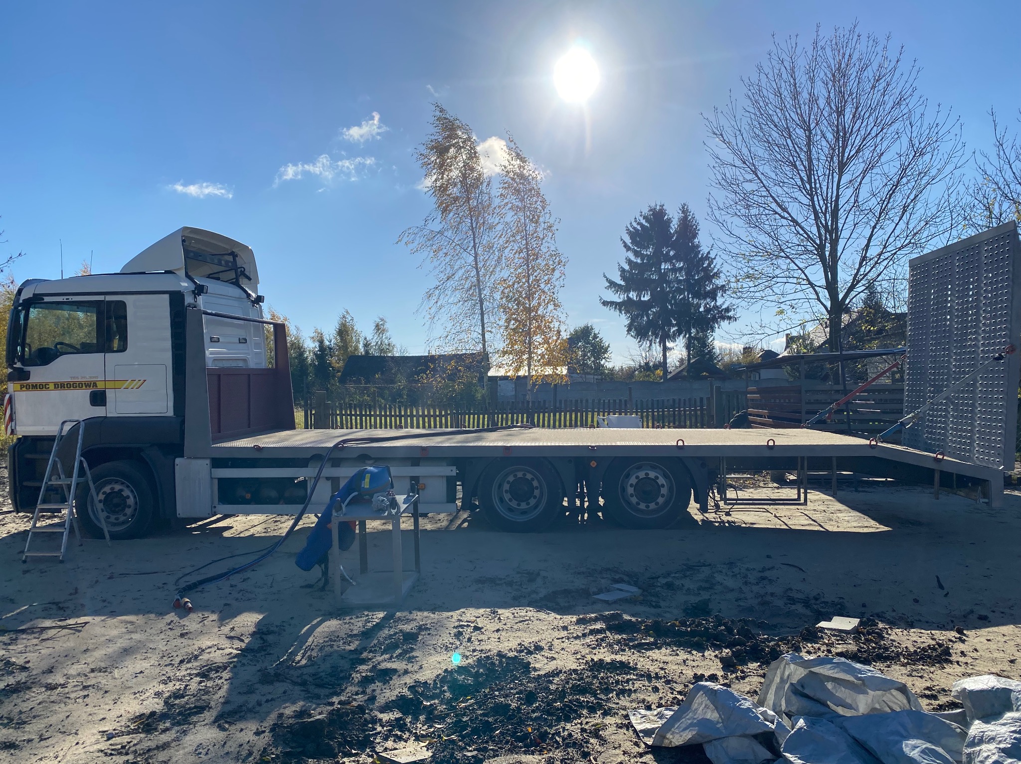Laweta pomocy drogowej z podnośnikiem hydraulicznym na tle błękitnego nieba i drzew. Sprzęt do czyszczenia na ziemi sugeruje przygotowanie do pracy.