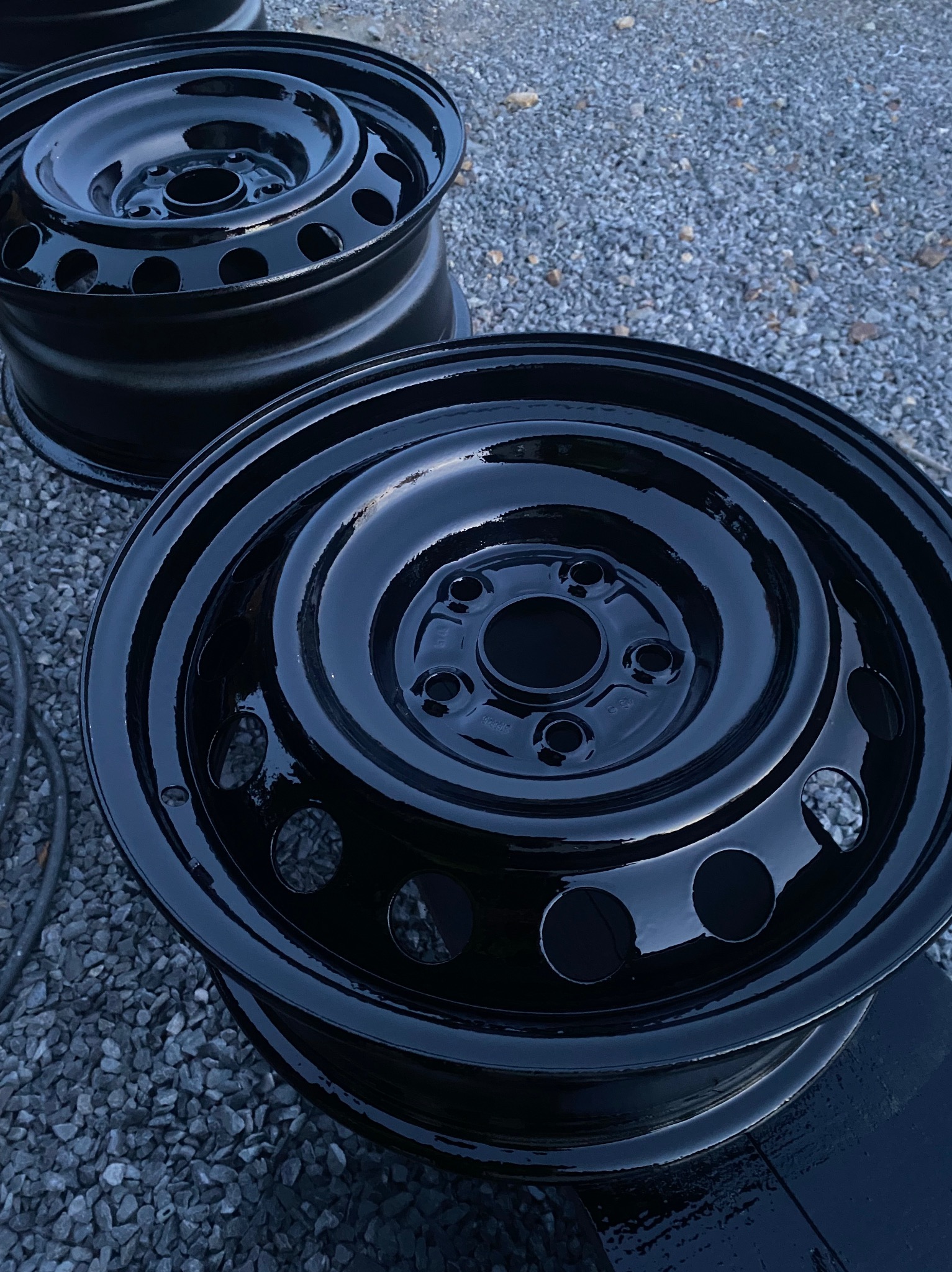 Błyszczące, czarne felgi stalowe po renowacji, ułożone na żwirowym podłożu. Widoczne detale odnowionej powierzchni i odbicia światła.