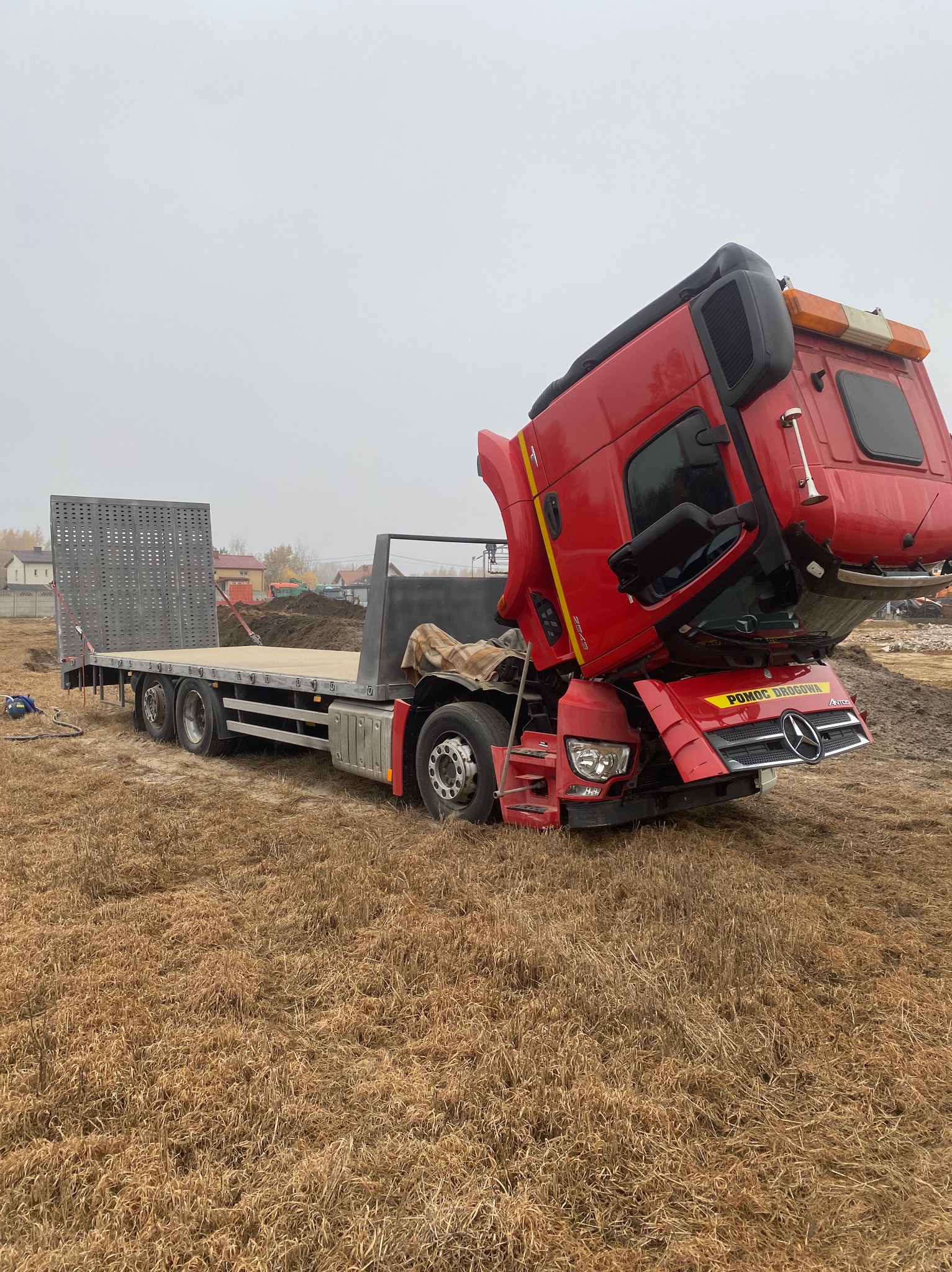 Czerwona ciężarówka z podniesioną kabiną na polu, widoczny napis 'Pomoc Drogowa' na masce. Platforma transportowa z tyłu. Pochmurne niebo w tle.