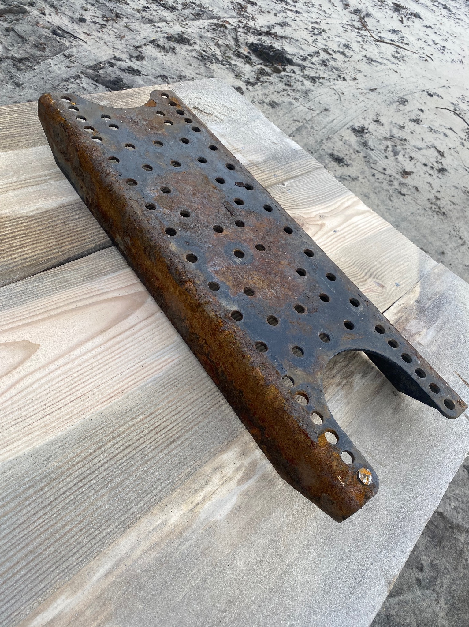 Skorodowana, metalowa platforma z otworami, leżąca na drewnianej powierzchni. Widoczne ślady rdzy i zużycia. Tło stanowi piaszczysta powierzchnia.