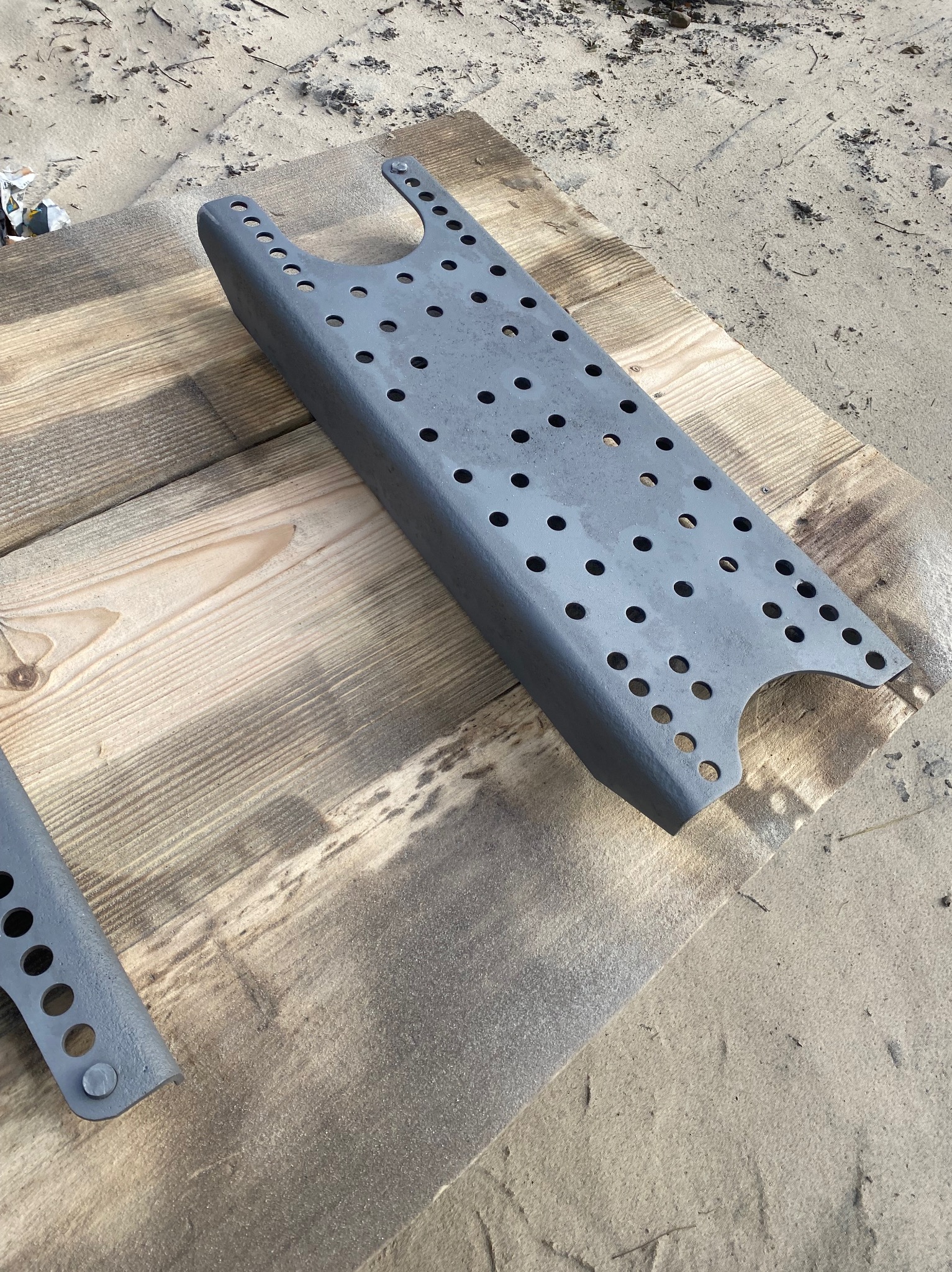 Szara, metalowa konstrukcja z otworami, leżąca na drewnianych deskach z piaskiem w tle. Element po zabezpieczeniu antykorozyjnym, gotowy do dalszego montażu.