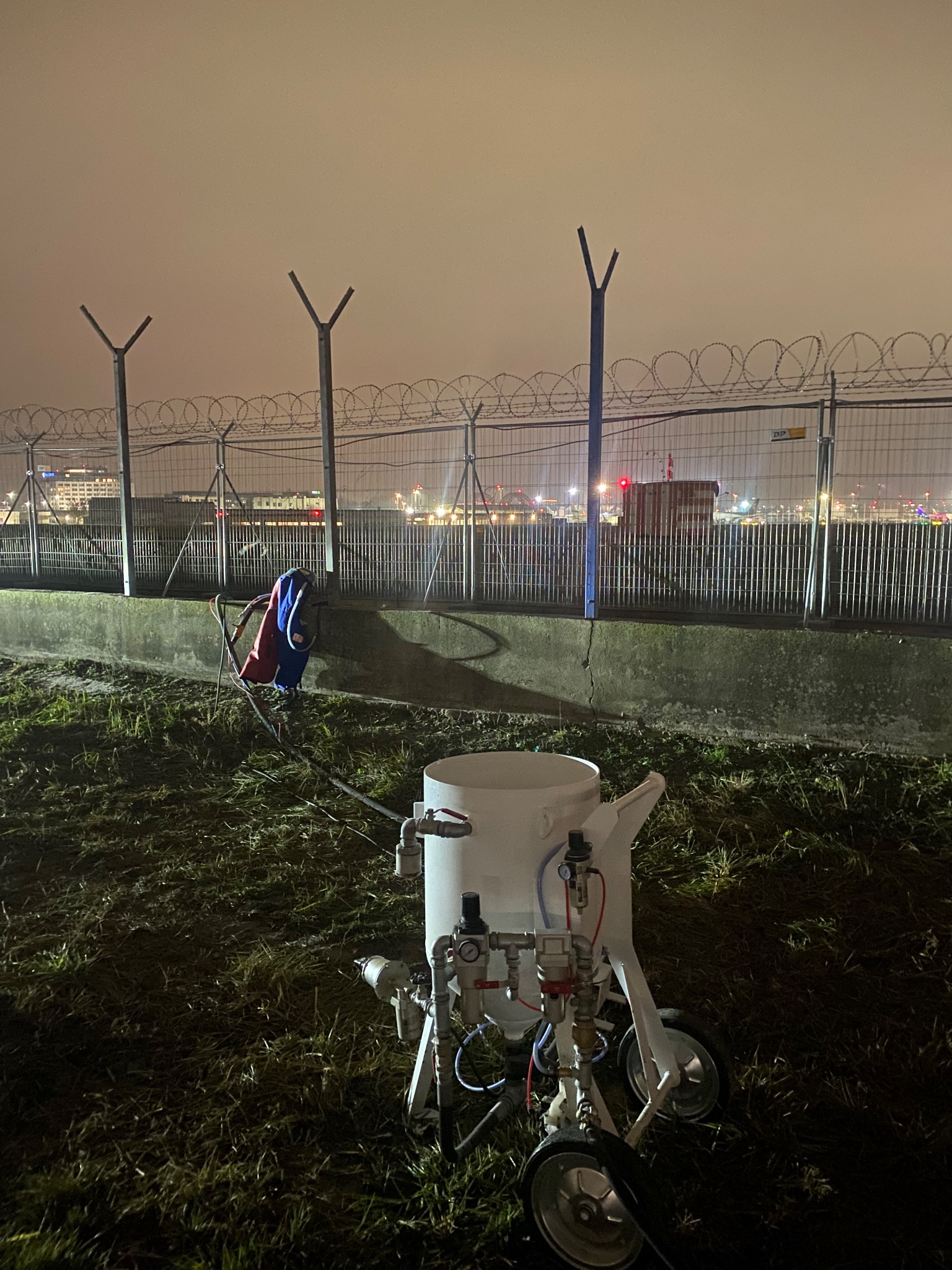 Mobilna piaskarka na trawie, w tle ogrodzenie z drutem kolczastym i budynki w oddali, nocne oświetlenie. Sprzęt do czyszczenia powierzchni w warunkach zewnętrznych.