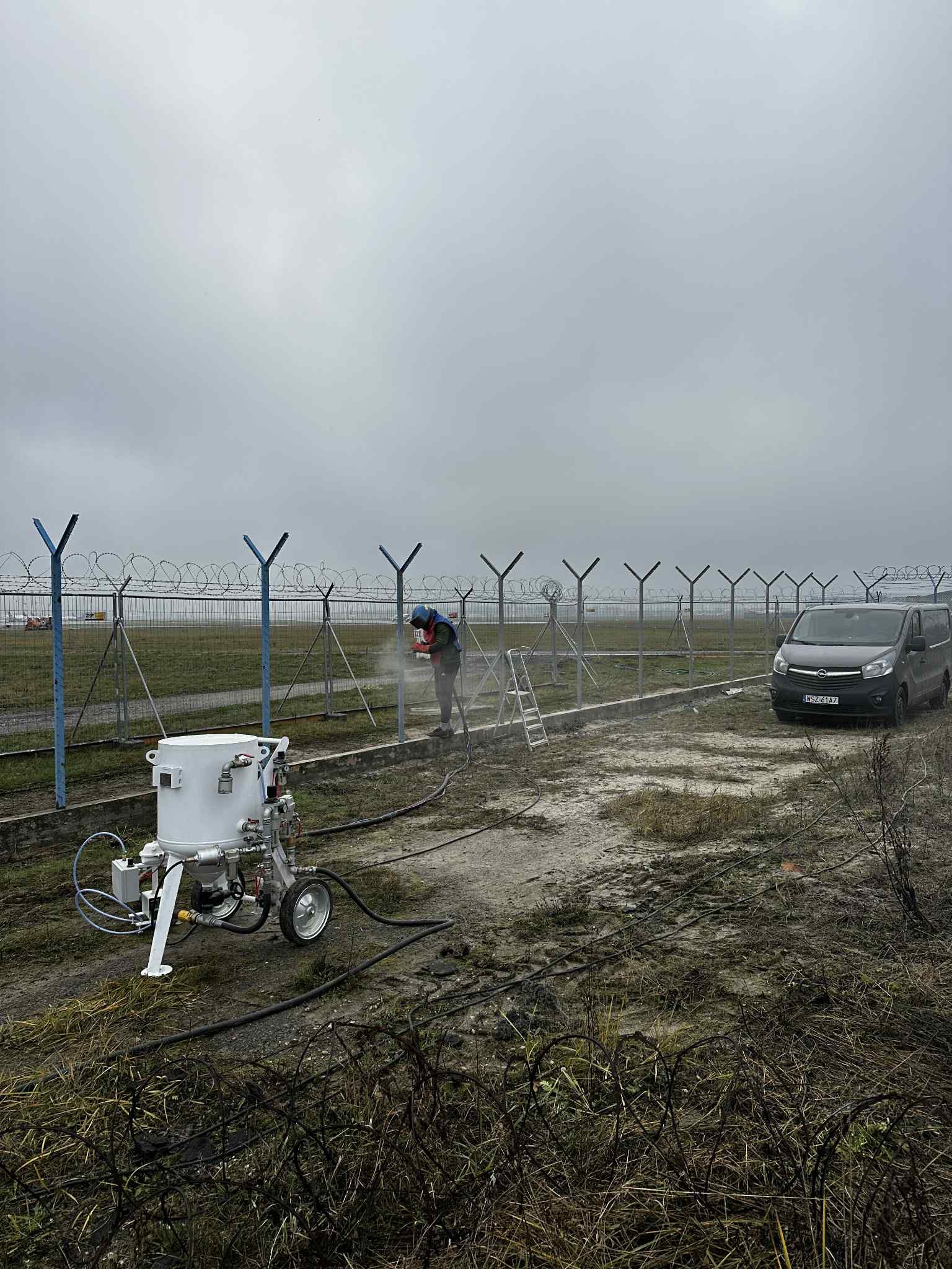 Pracownik w kombinezonie czyści metalowe ogrodzenie z drutem kolczastym za pomocą mobilnego urządzenia. W tle szary van i pochmurne niebo.