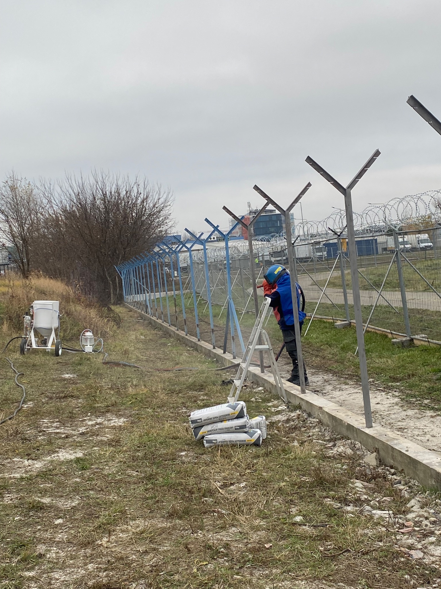 Pracownik w kombinezonie ochronnym czyści metalowe ogrodzenie z drutem kolczastym za pomocą specjalistycznego sprzętu do piaskowania. Widoczne worki z piaskiem.