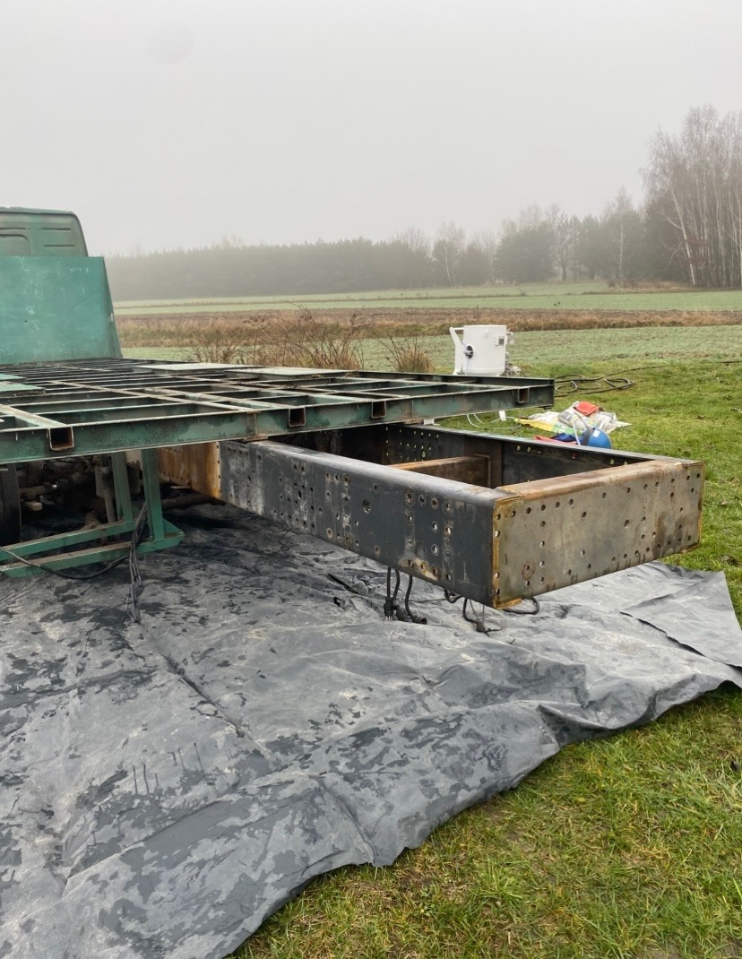 Oczyszczona rama pojazdu na zewnątrz, widoczne ślady korozji i przygotowania do dalszej obróbki. Tło: łąka i mglisty las.