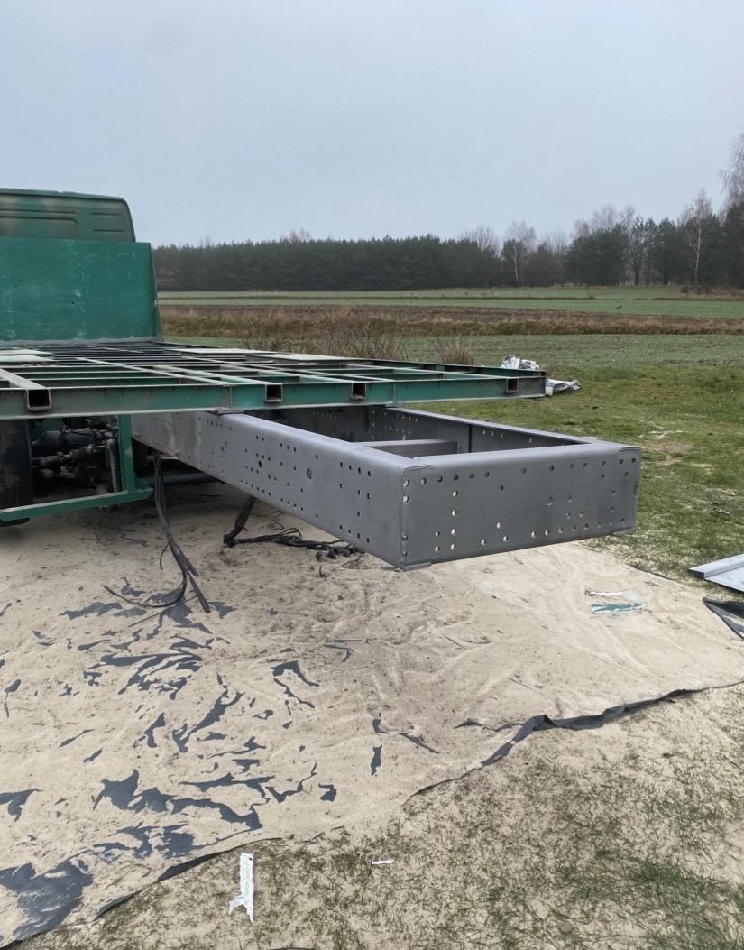 Szara rama pojazdu z otworami, częściowo pomalowana na zielono, na tle łąki i lasu. Prace konserwacyjne na zewnątrz, na rozłożonej plandece.