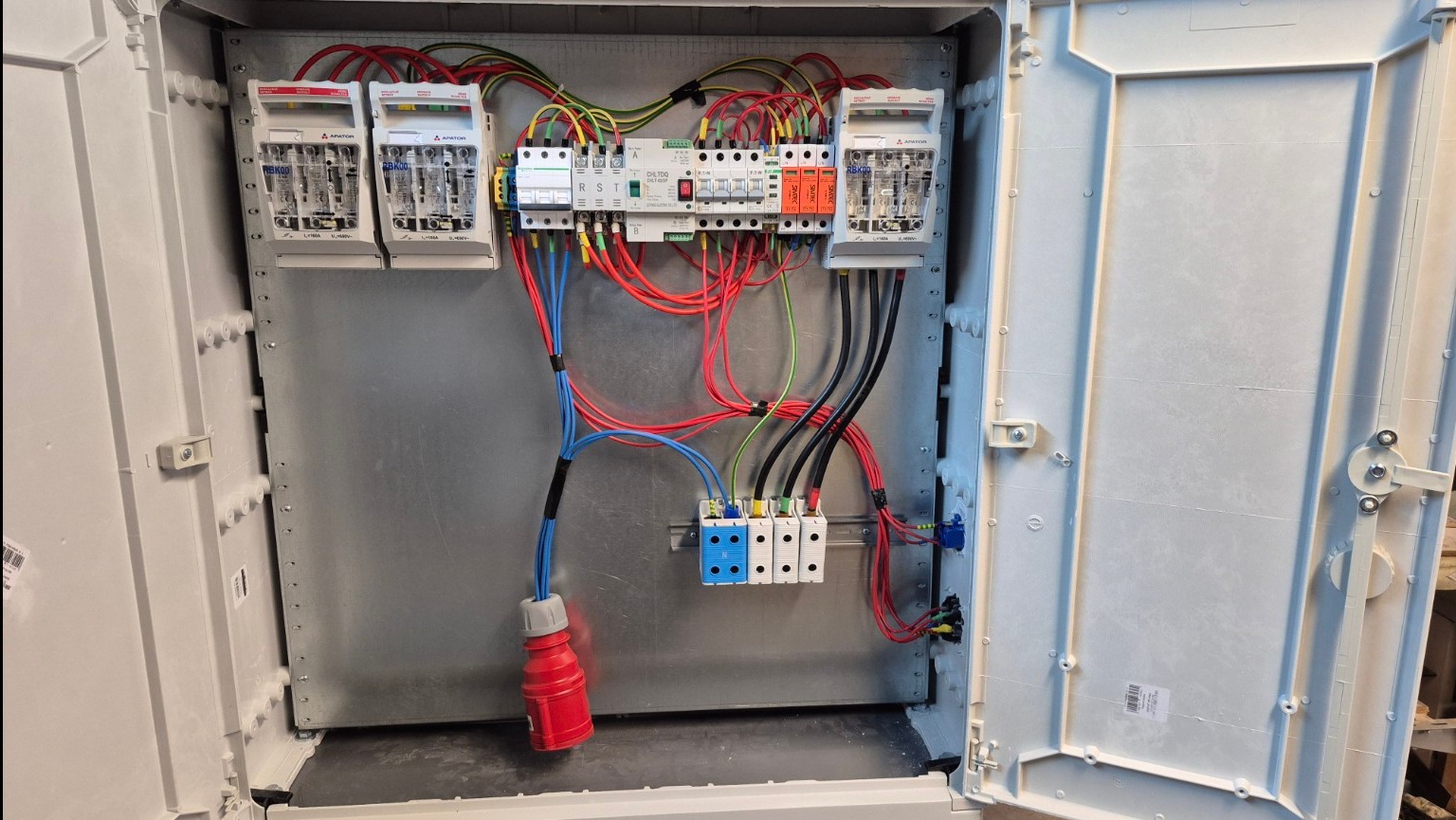 Otwarta skrzynka elektryczna z bezpiecznikami, przewodami w kolorach: czerwonym, niebieskim, czarnym i zielono-żółtym. Widoczny czerwony wtyk. Metalowa płyta montażowa.