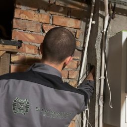 SIGMATOM SPÓŁKA Z OGRANICZONĄ ODPOWIEDZIALNOŚCIĄ - Elektryk w szarym uniformie montuje białe rury instalacji elektrycznej na ceglanej ścianie. Widoczne kable, skrzynka elektryczna i metalowa konstrukcja pod sufitem.
