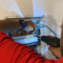 SIGMATOM SPÓŁKA Z OGRANICZONĄ ODPOWIEDZIALNOŚCIĄ - Elektryk w trakcie montażu bezpieczników w skrzynce elektrycznej, używając wkrętarki. Widoczne przewody i elementy instalacji. Ściana z widocznymi pęknięciami.