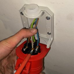 SIGMATOM SPÓŁKA Z OGRANICZONĄ ODPOWIEDZIALNOŚCIĄ - Elektryk podłącza przewody do czerwonego gniazda elektrycznego w białej obudowie, używając śrubokręta. Widoczne przewody w różnych kolorach.