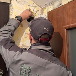 SIGMATOM SPÓŁKA Z OGRANICZONĄ ODPOWIEDZIALNOŚCIĄ - Elektryk w trakcie pracy nad instalacją elektryczną wewnątrz budynku. Widoczne przewody, ściana z uszkodzonym tynkiem i fragment firmowego ubrania.