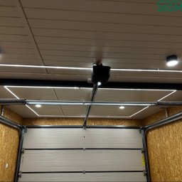 SIGMATOM SPÓŁKA Z OGRANICZONĄ ODPOWIEDZIALNOŚCIĄ - Zamknięta brama garażowa segmentowa z automatyką i oświetleniem LED. Widoczne sprężyny i mechanizm otwierania. Ściany wyłożone płytami OSB.