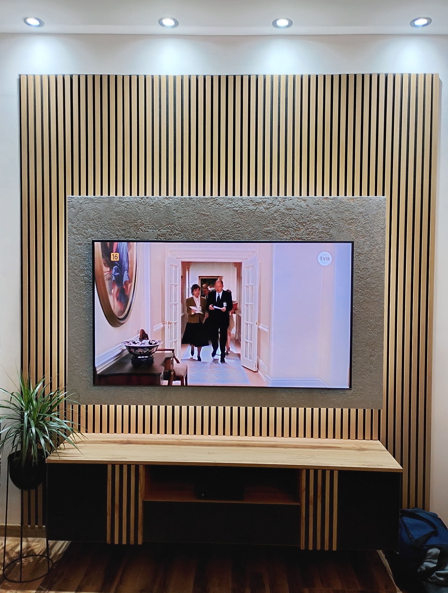 Nowoczesna ściana TV z drewnianymi lamelami i szarą ramą, podświetlona punktowo. Na szafce RTV stoi roślina doniczkowa. Widoczny fragment podłogi.