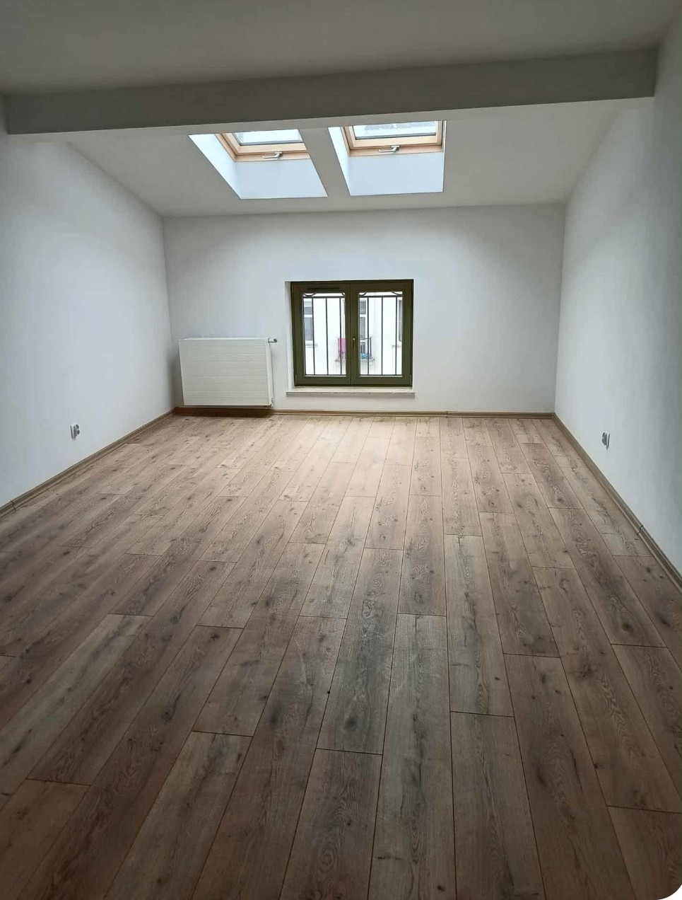 Jasne wnętrze po remoncie: drewniana podłoga, białe ściany, okno z kratą i dwa okna dachowe. Minimalistyczny grzejnik przy ścianie.