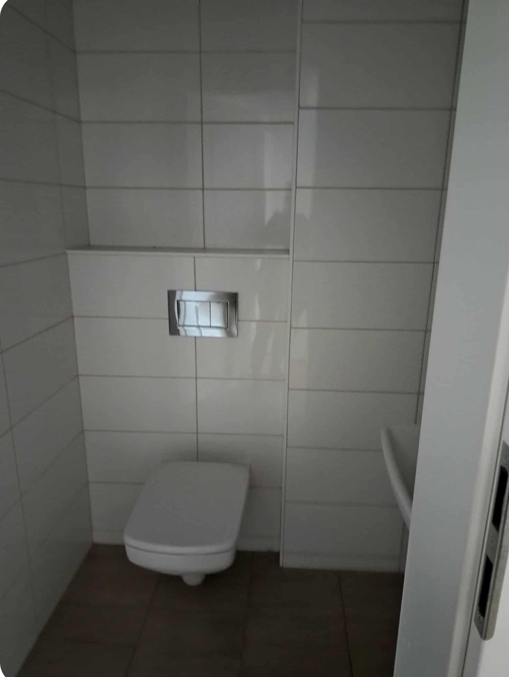 Nowoczesna toaleta z białymi płytkami i podwieszaną muszlą klozetową. Widoczny chromowany przycisk spłukiwania i ciemna podłoga. Minimalistyczny design.