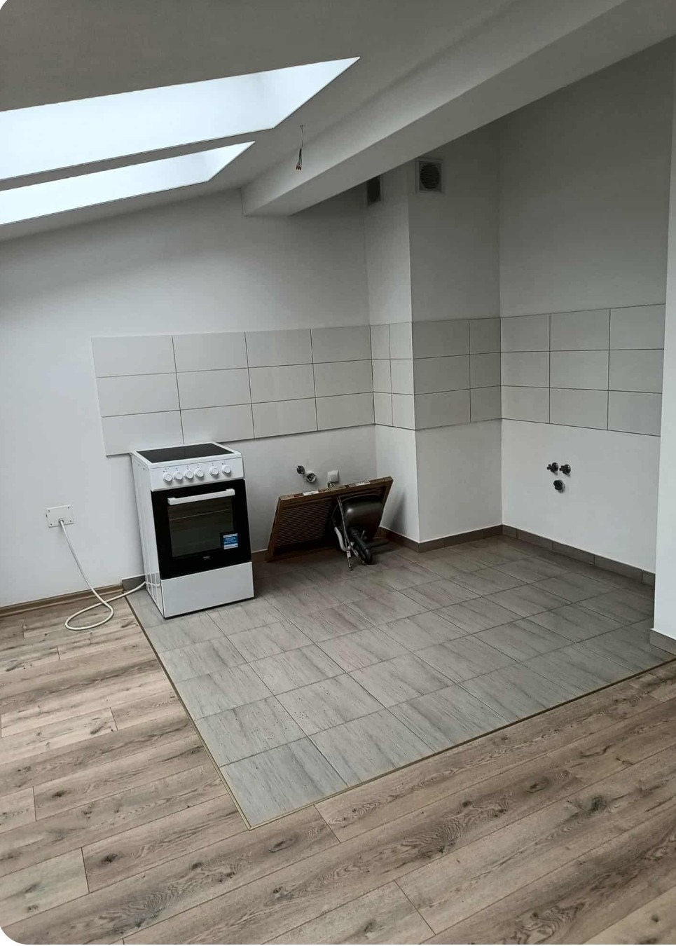 Wykończona kuchnia z płytkami na ścianie i podłodze, widoczna kuchenka. Poddaszowe okno doświetla wnętrze. Część podłogi wyłożona panelami.