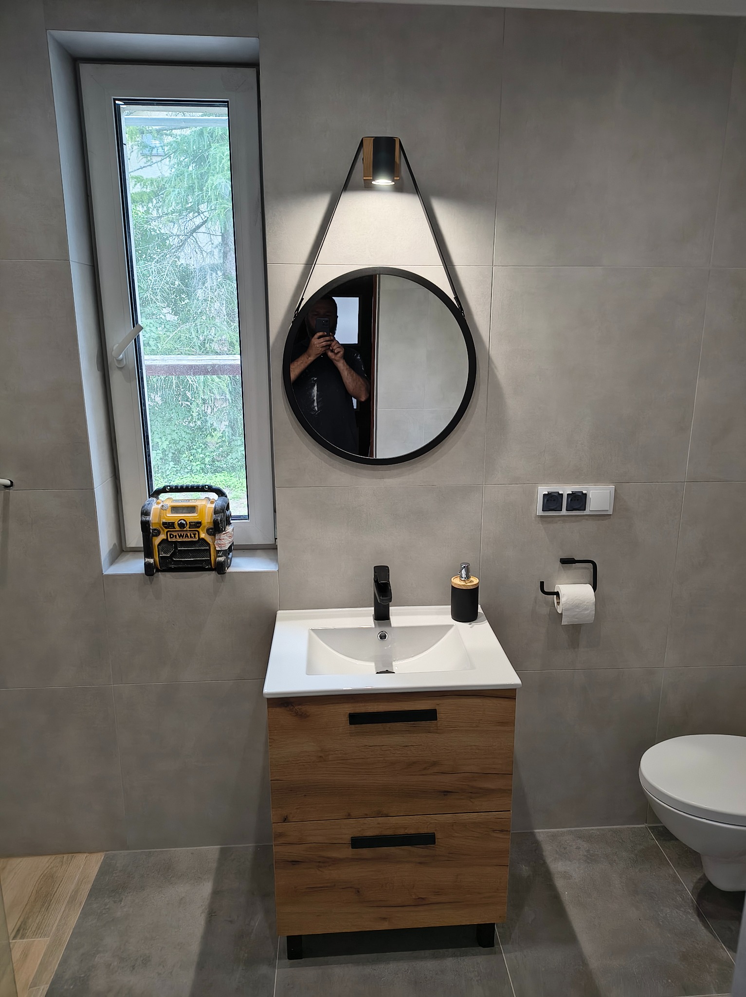 Nowoczesna łazienka z szarymi płytkami, drewnianą szafką pod umywalką i okrągłym lustrem z oświetleniem. Na parapecie żółta ładowarka akumulatorów.