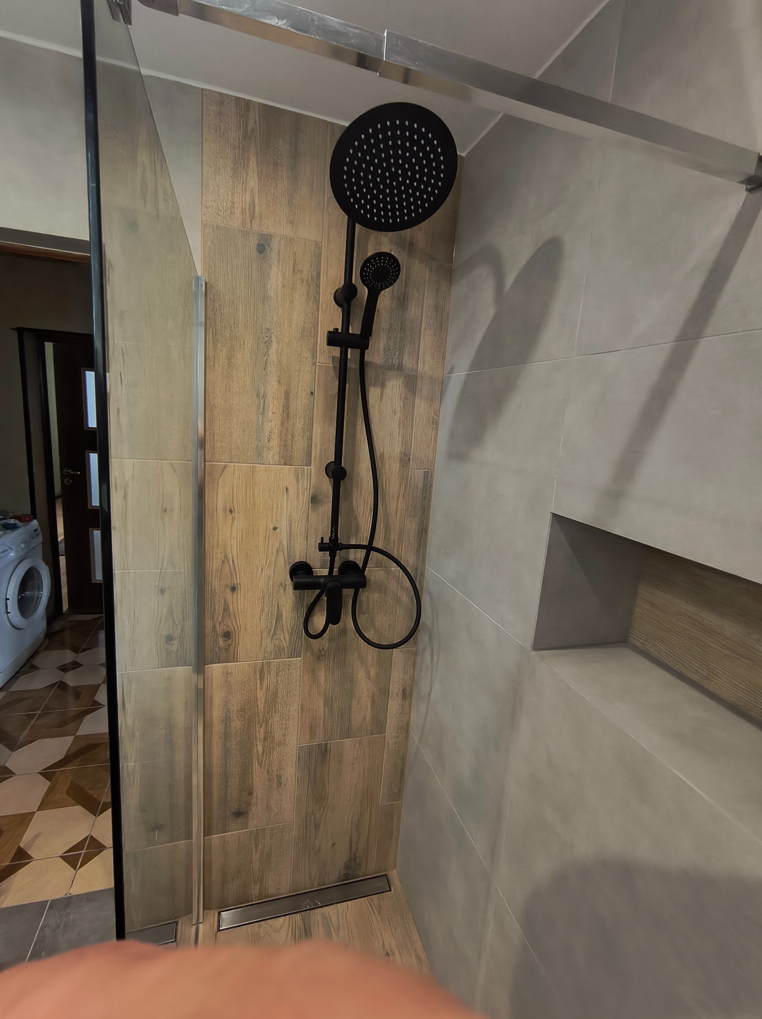 Nowoczesna kabina prysznicowa z czarną armaturą i odpływem liniowym. Ściany wyłożone płytkami imitującymi drewno i szarym gresem. Widoczna pralka w tle.