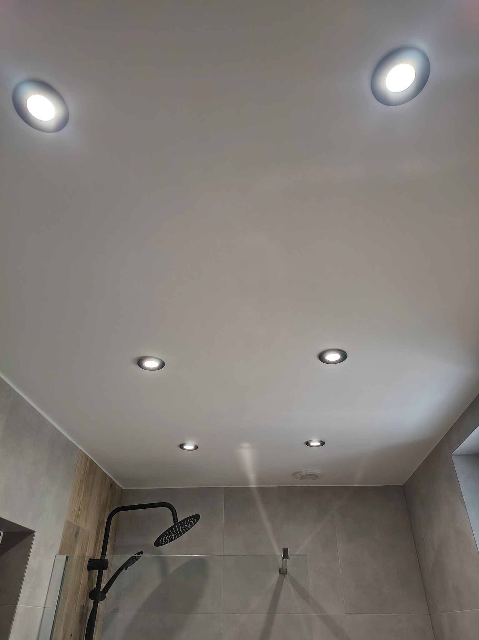 Nowoczesna łazienka z szarą glazurą, czarną armaturą i oświetleniem punktowym w suficie podwieszanym. Minimalistyczny design, widok z dołu.