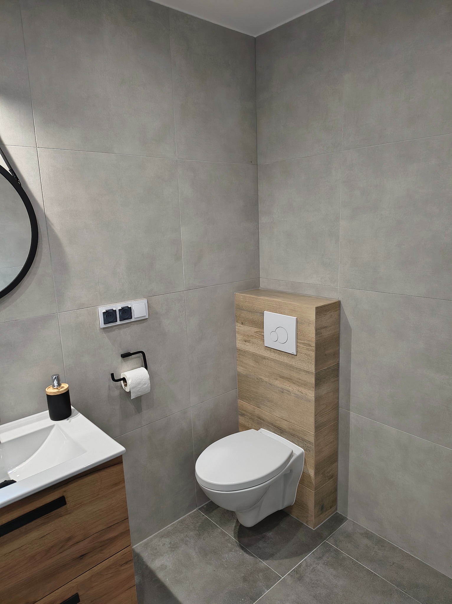 Nowoczesna łazienka z szarymi płytkami, podwieszaną toaletą z drewnianą obudową i umywalką z szafką w stylu drewna. Minimalistyczny design, czarne akcenty.