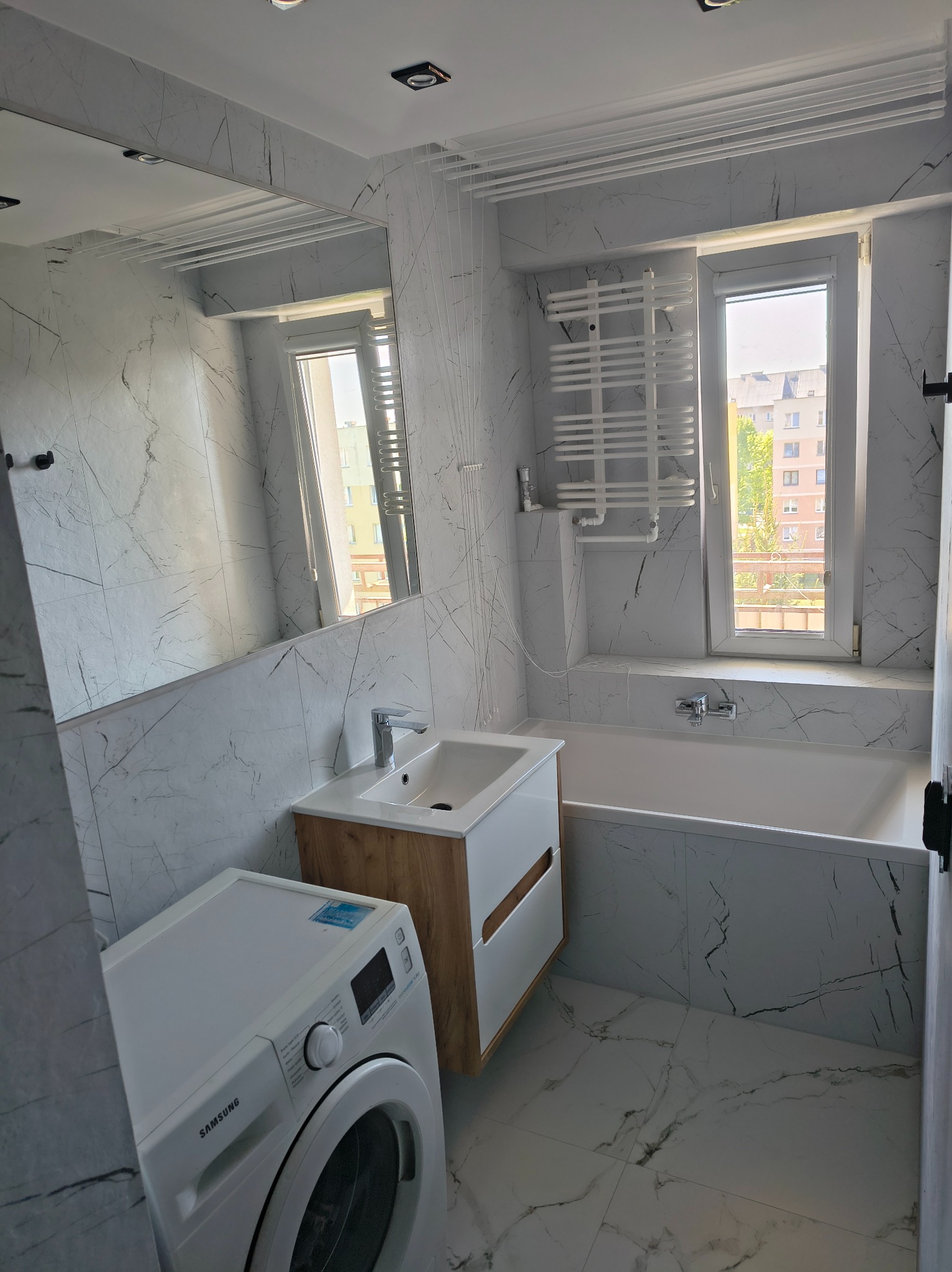 Nowoczesna łazienka z marmurowymi płytkami, drewnianą szafką pod umywalką i pralką Samsung. Widok na okno z suszarką sufitową.