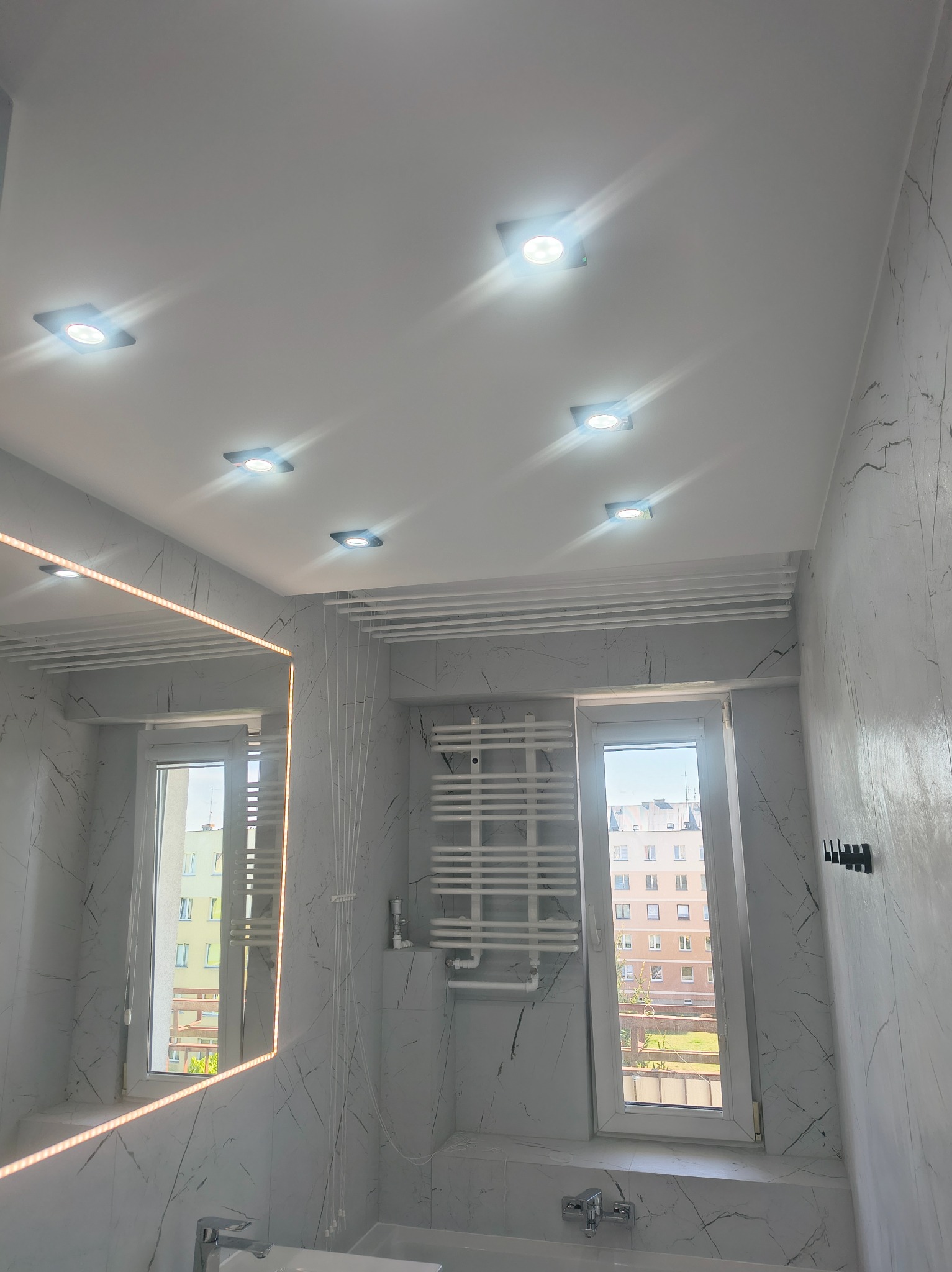 Wykończona łazienka w bieli i szarości z marmurkową teksturą, oświetleniem punktowym i lustrem z podświetleniem LED. Widoczny grzejnik drabinkowy i okno.