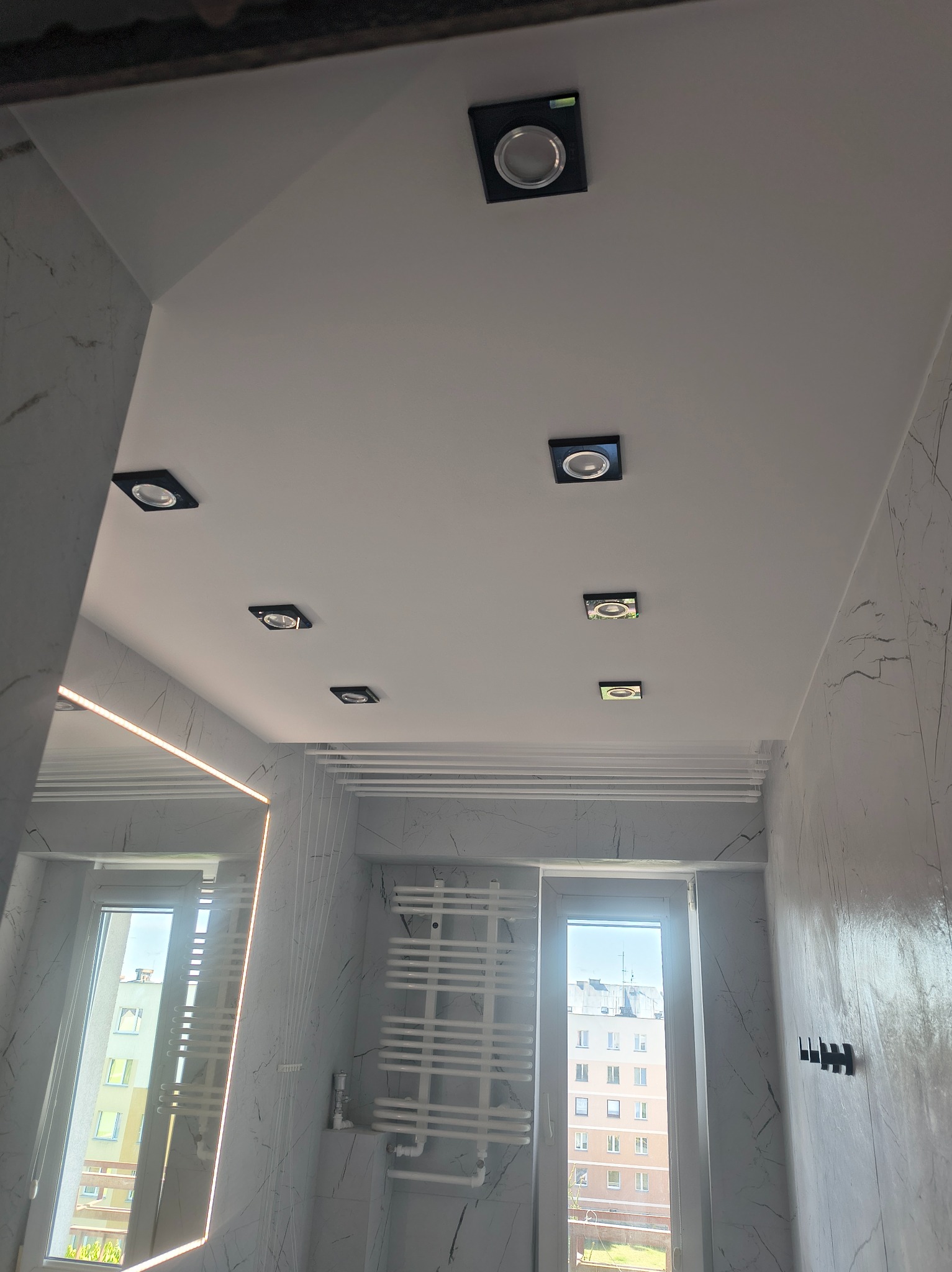 Biały sufit w łazience z zamontowanymi kwadratowymi lampami punktowymi. Na ścianach płytki imitujące marmur. Widoczne okno i grzejnik drabinkowy.