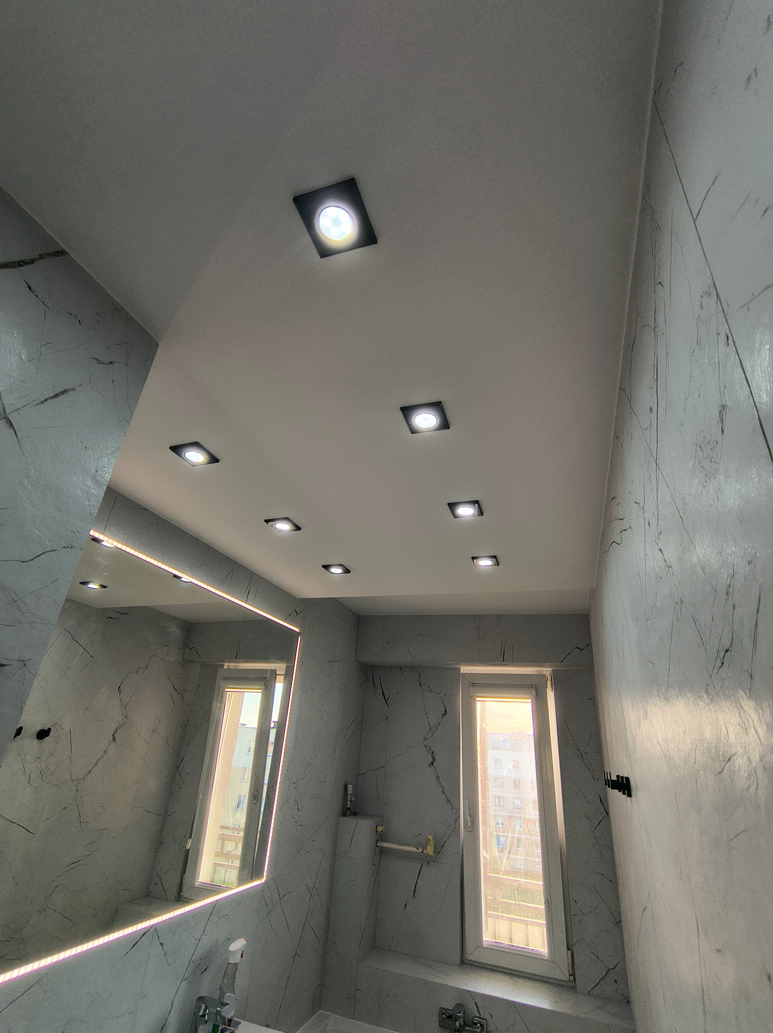 Wykończona łazienka w szarościach z marmurkową teksturą na ścianach, oświetleniem punktowym w suficie i lustrem z podświetleniem LED. Widok na okno.